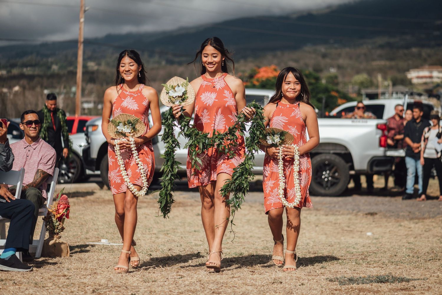 Three bridesmaids in matching floral dresses walk outdoors carrying bouquets at a Hawaiian wedding ceremony.