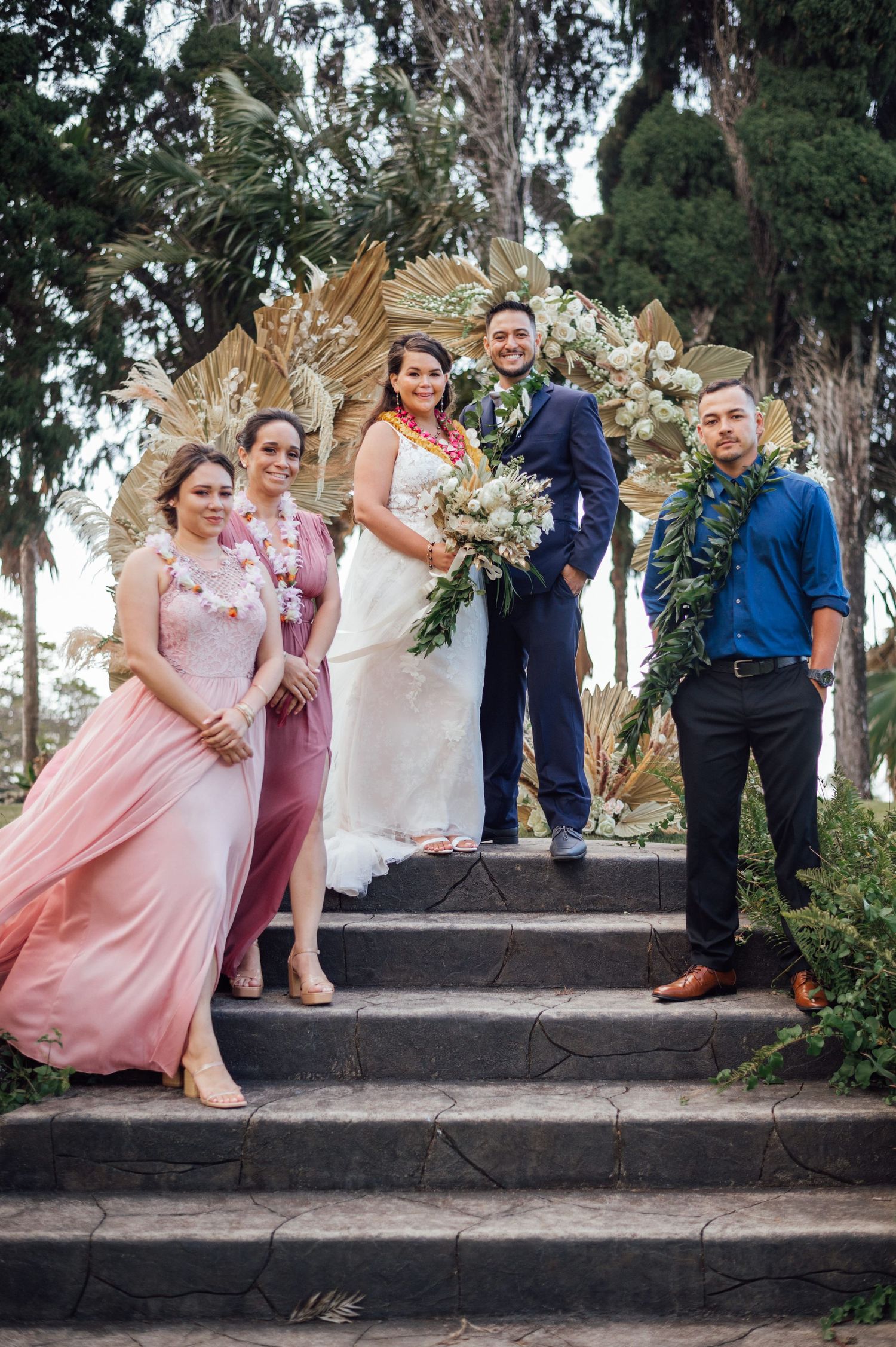 Wedding party poses on stone steps with floral arch backdrop, bride in white gown, groom in navy suit, bridesmaids in pink.