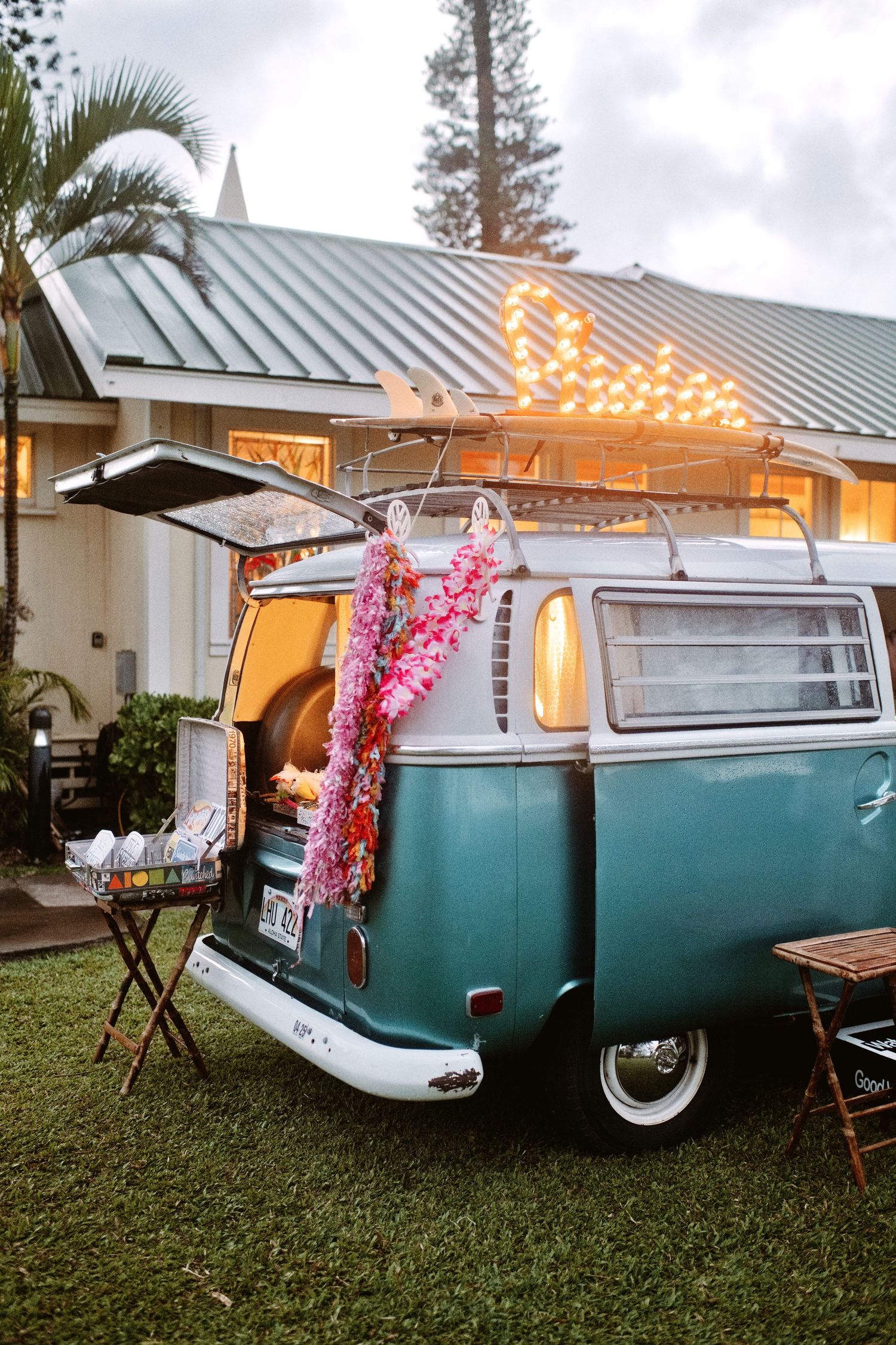 Vintage teal VW bus decorated with fairy lights and floral garlands, set up as an outdoor event bar on green lawn.