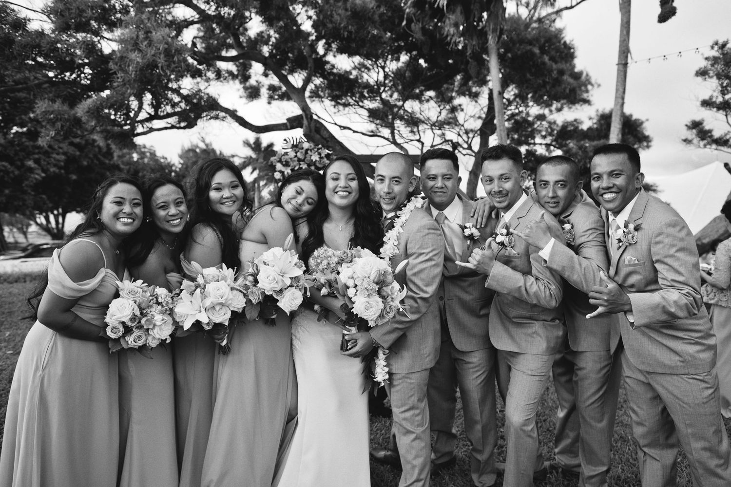 Black and white wedding party photo with bride, groom, bridesmaids, and groomsmen laughing outdoors.