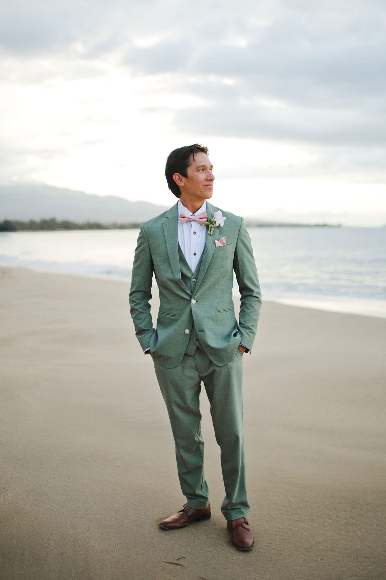 Groom in mint green three-piece suit with boutonniere stands on sandy beach with ocean backdrop.