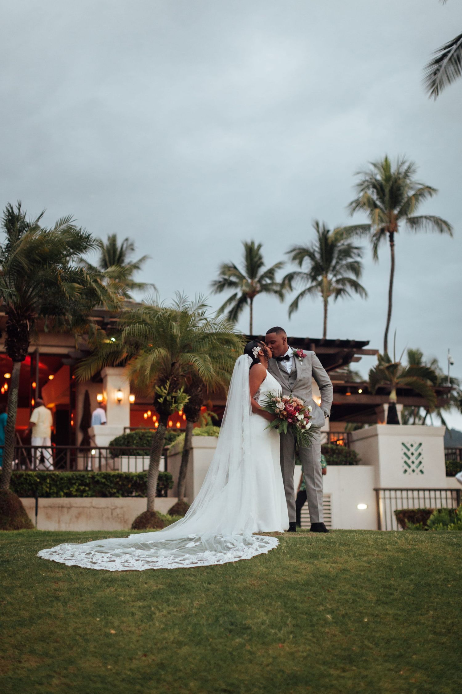 Bride and groom share a romantic kiss on a lush lawn with palm trees and ocean views at a tropical wedding venue.