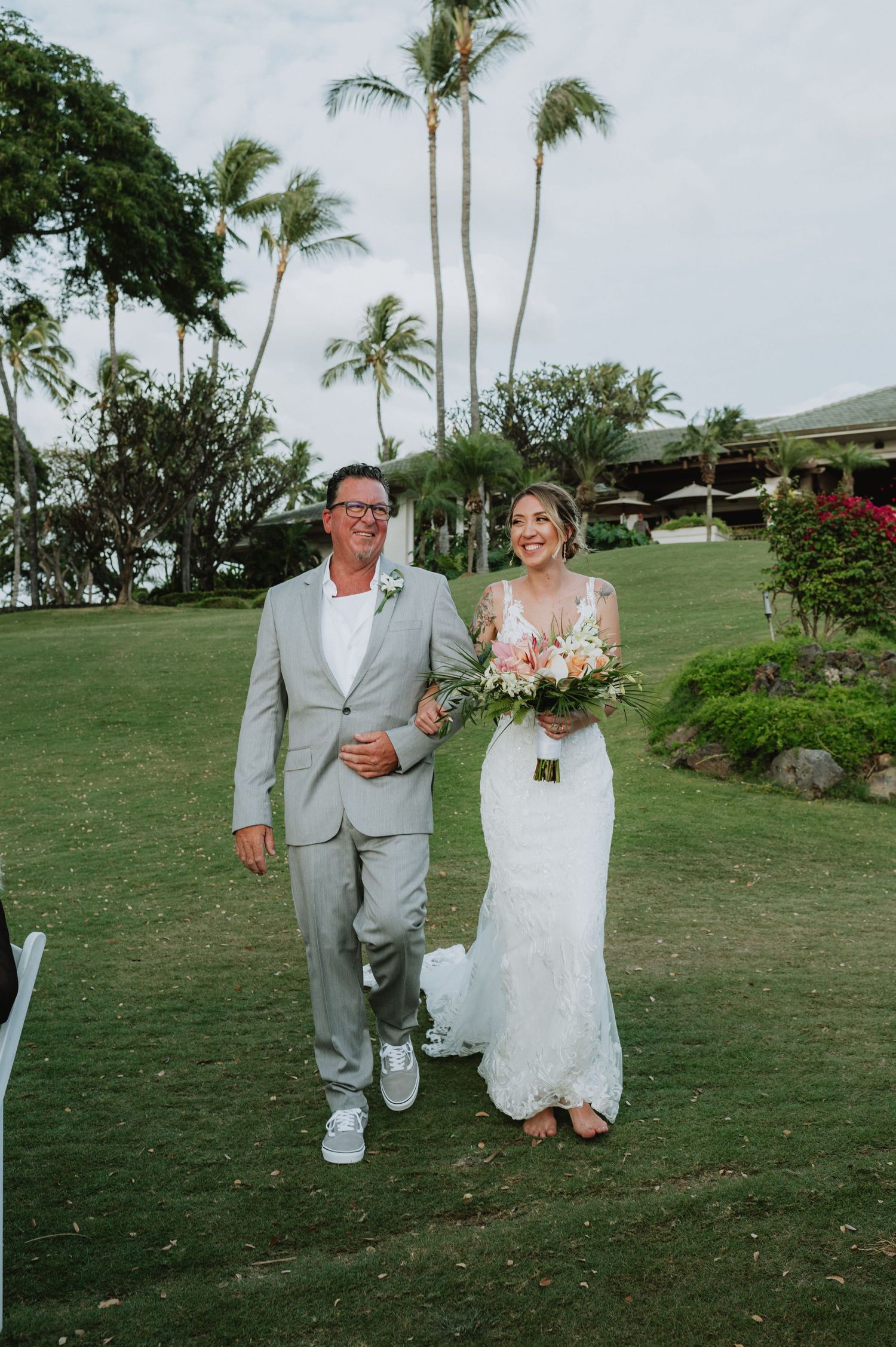 Bride in lace gown holding floral bouquet walks down outdoor aisle with man in gray suit at tropical Hawaii wedding.