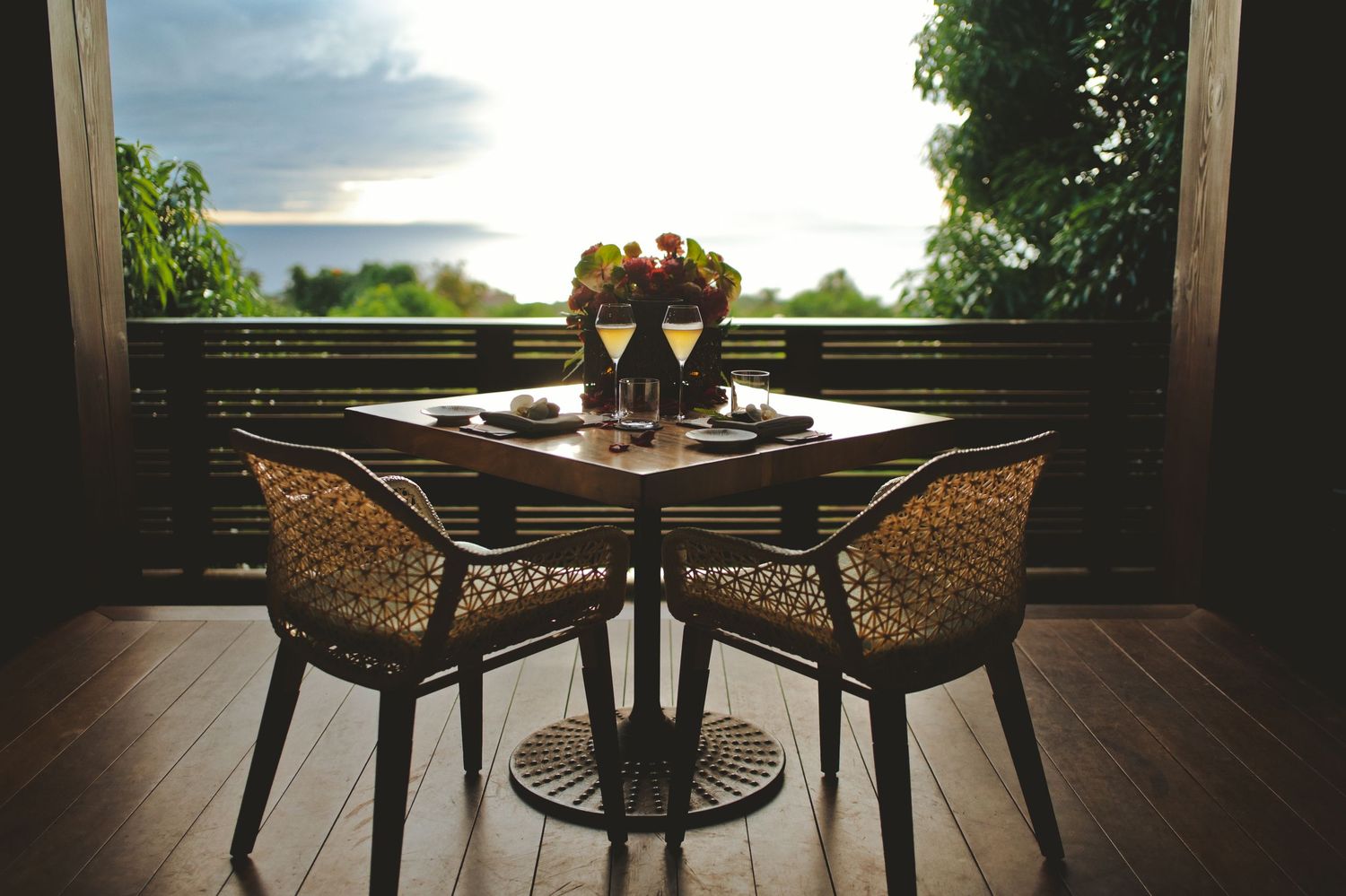 Romantic outdoor dining table with wicker chairs, wine glasses, and ocean sunset view from a tropical resort balcony.