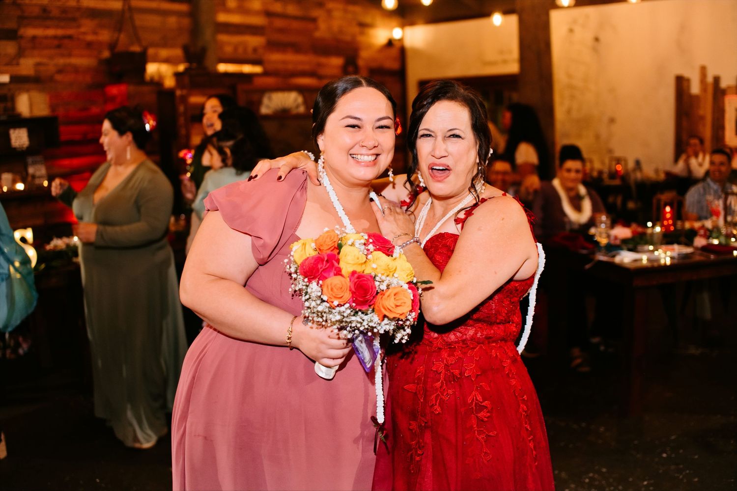 Two women in formal dresses smiling and posing together at a wedding reception, one holding a colorful bouquet.