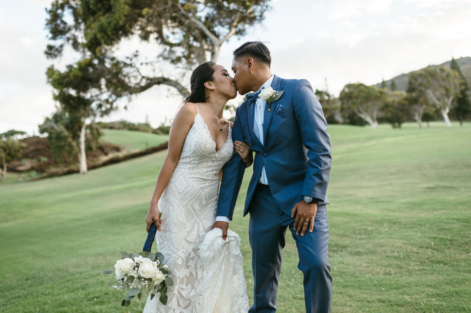 Elegant wedding couple poses on grassy hillside in formal attire.