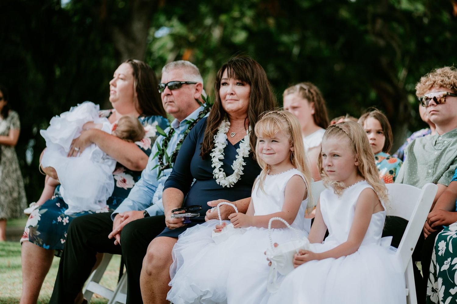 Wedding guests seated in white chairs watch an outdoor ceremony under palm trees.
