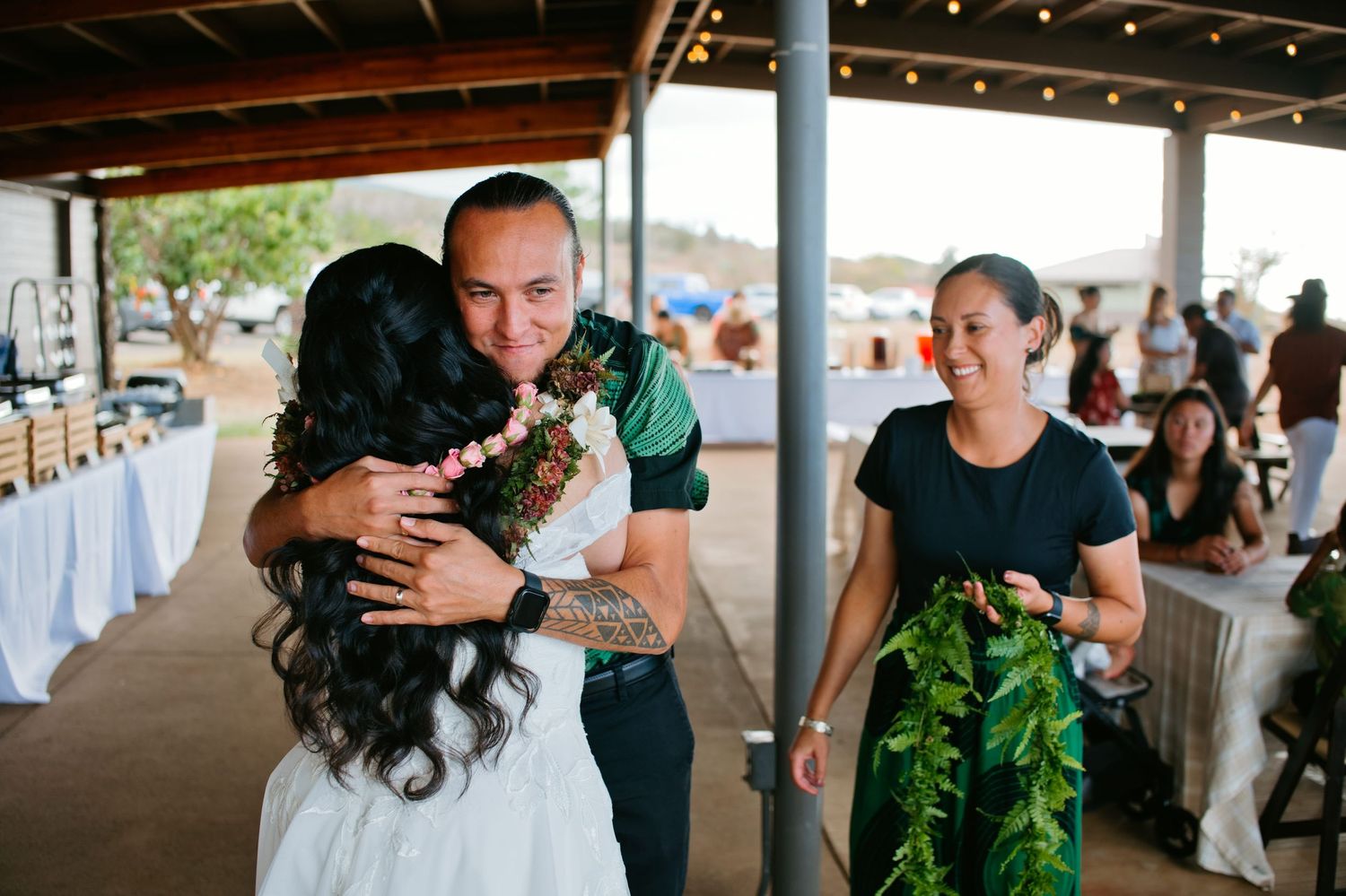 Couple embracing at outdoor Hawaiian wedding reception with floral leis and guests in background.