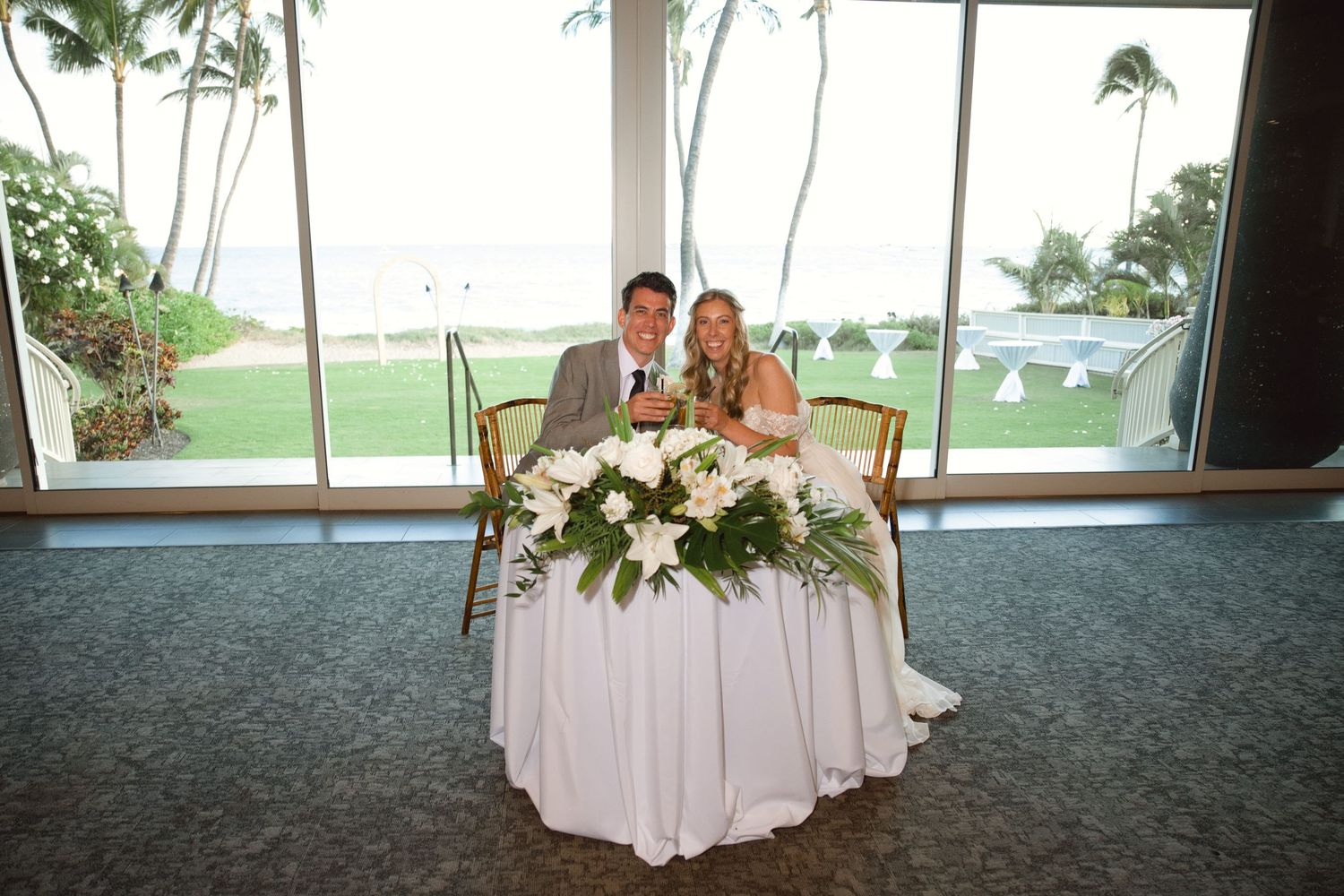 Bride and groom seated at sweetheart table with white floral centerpiece, ocean view backdrop at Hawaii wedding venue.