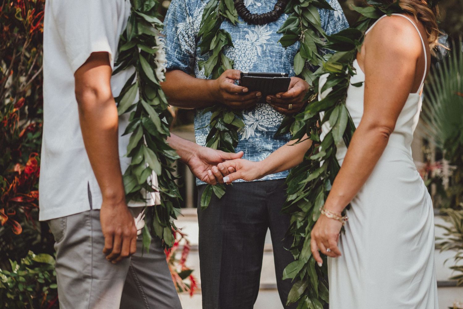 Couple holding hands during Hawaiian wedding ceremony with officiant wearing lei and blue floral shirt.