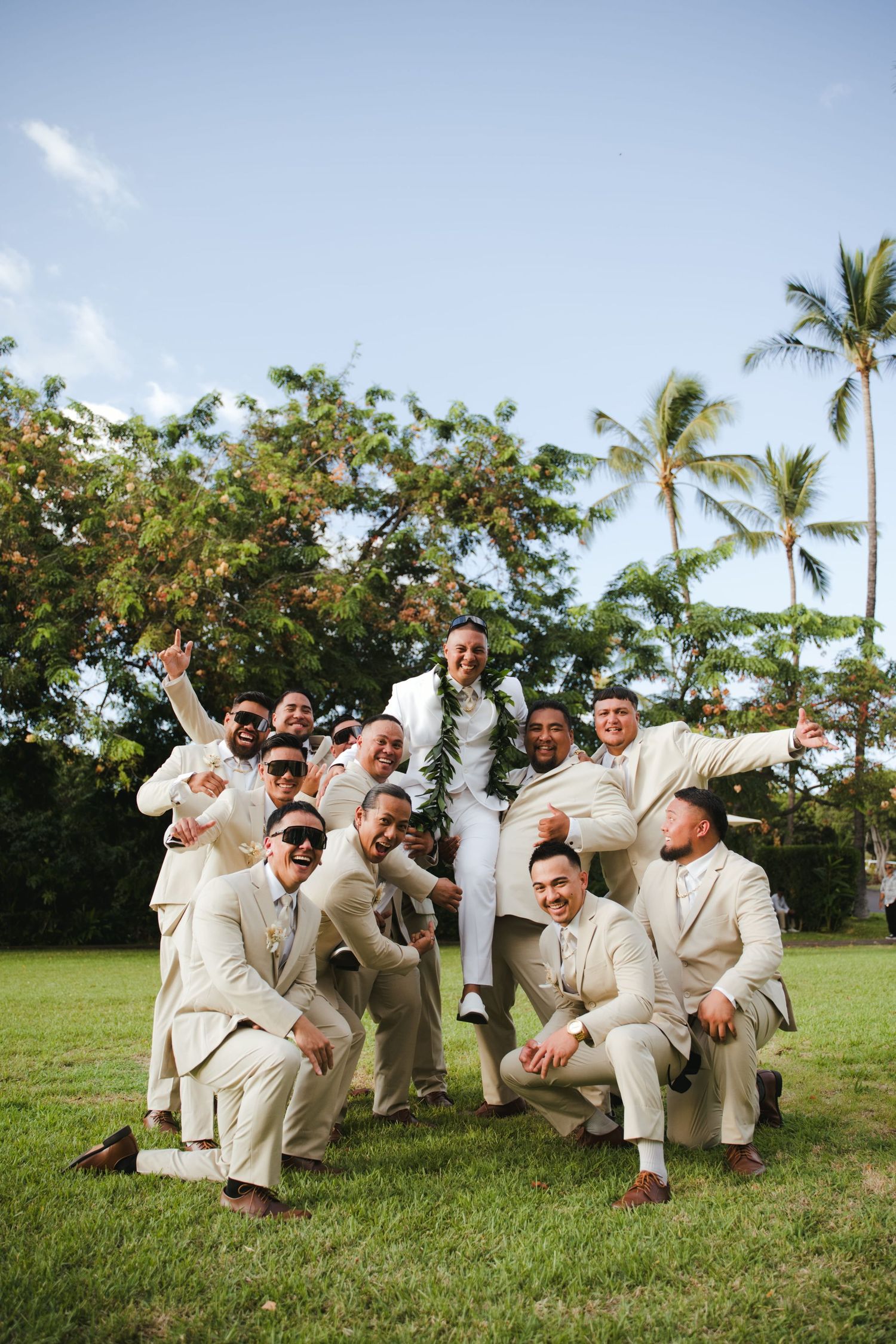 Groom lifted by groomsmen in tan suits on green lawn with palm trees at tropical Hawaii wedding.