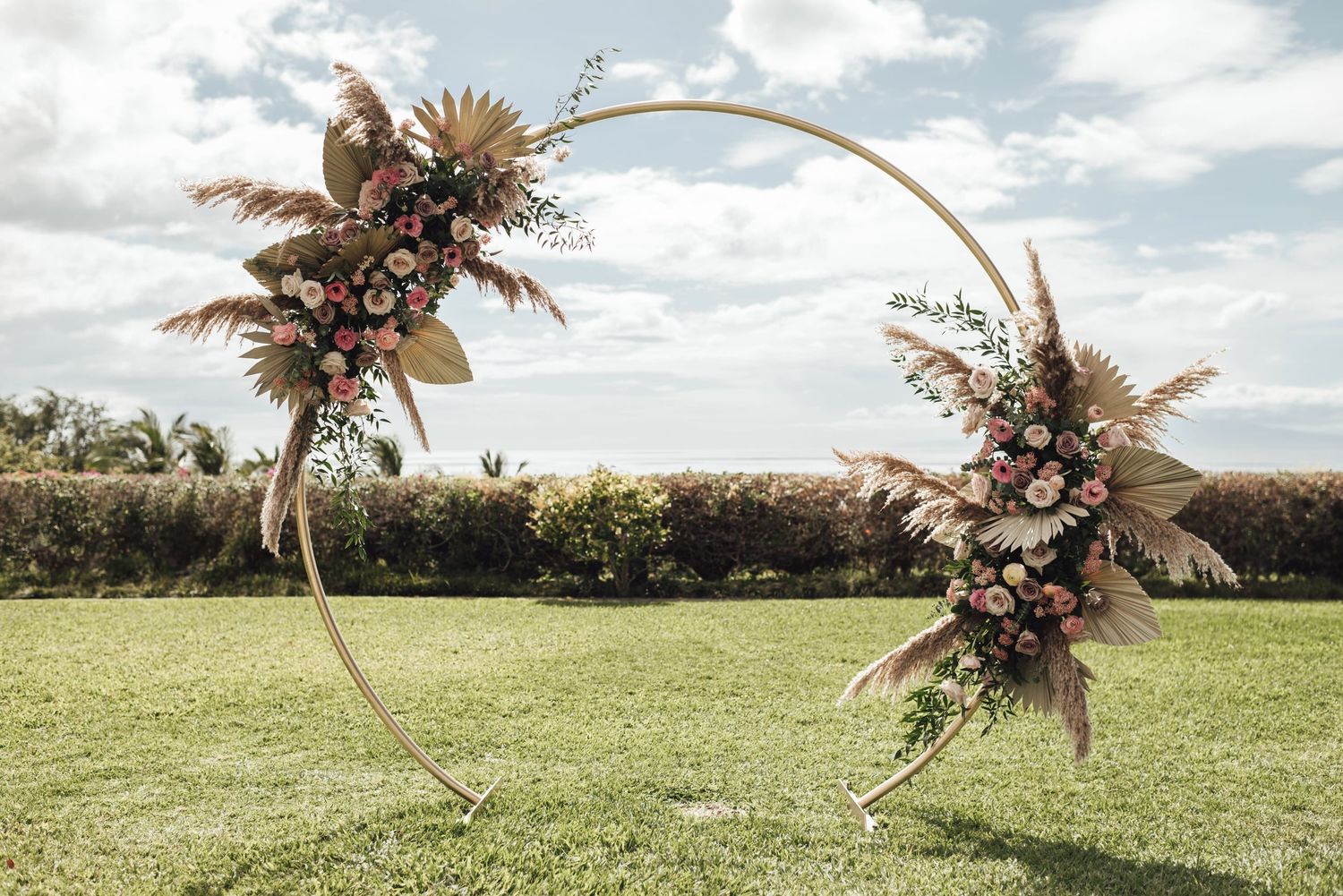 Circular gold wedding arch adorned with pampas grass and pink flowers on green lawn under cloudy sky.