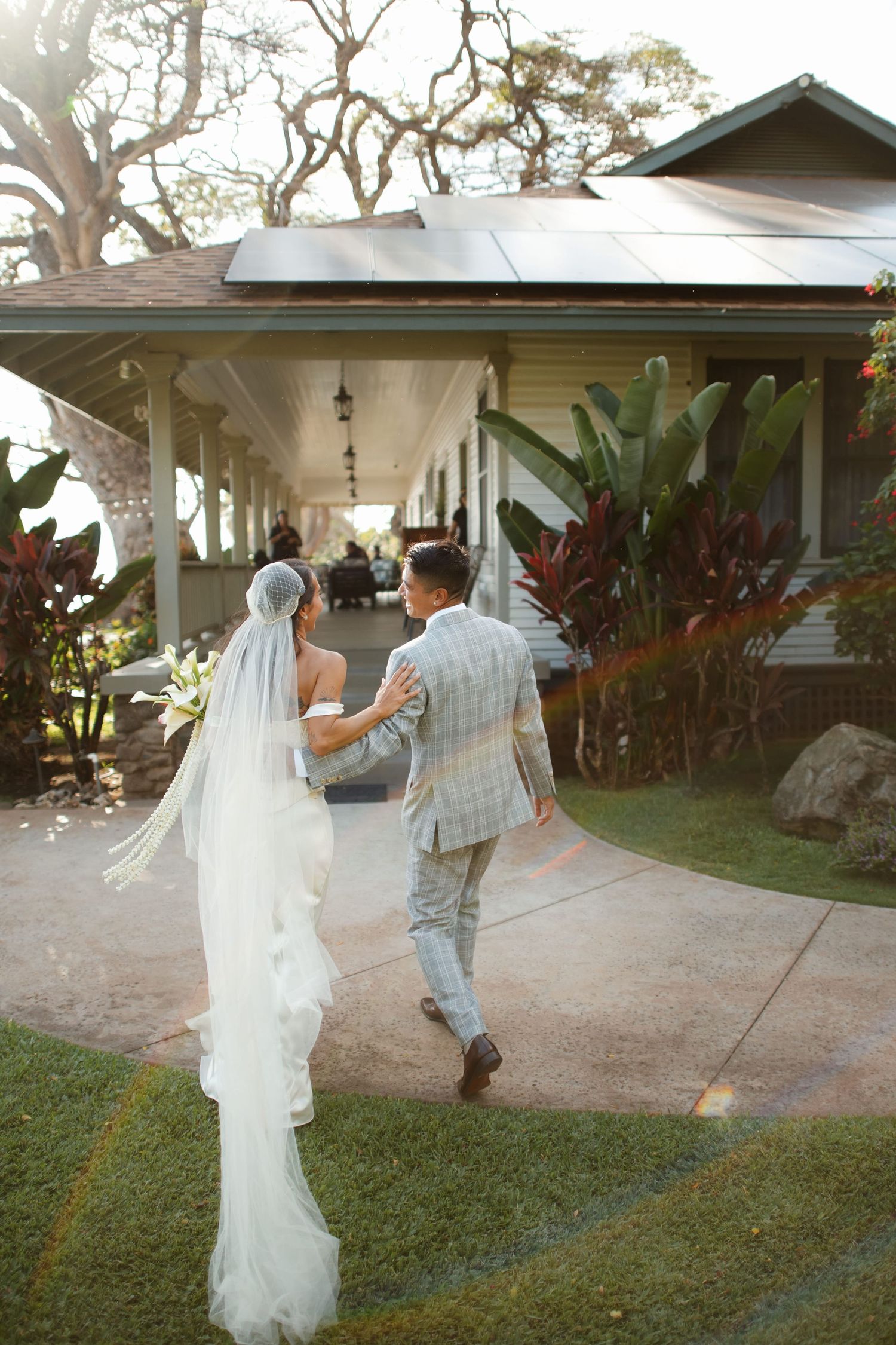 Bride in white gown and veil walks with groom in plaid suit toward tropical venue porch, viewed from behind.