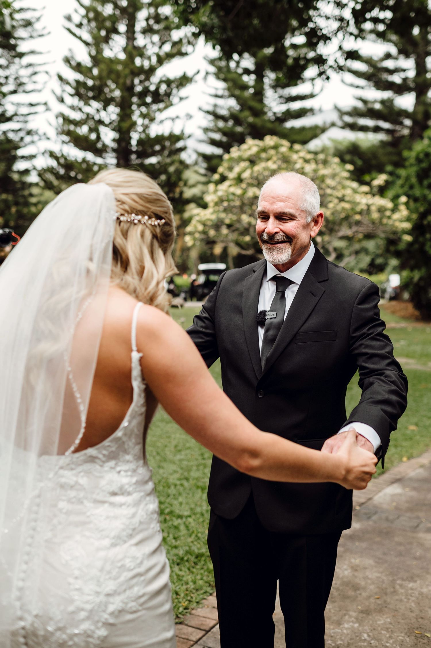 A smiling older man in a black suit holds hands with a bride in a white lace wedding dress and veil outdoors.