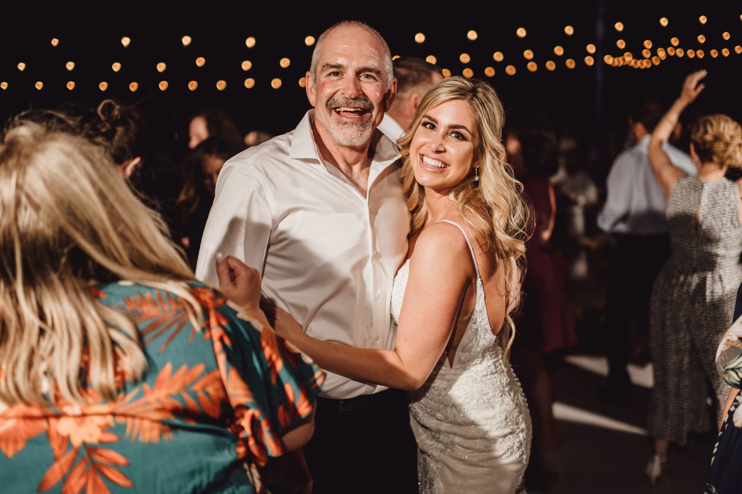 Bride and older man share a joyful dance together at a wedding reception under warm string lights.