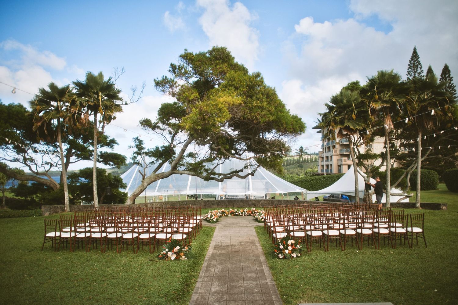 Outdoor wedding ceremony setup with white tent, wooden chairs, and floral aisle on lush green lawn surrounded by tropical trees.