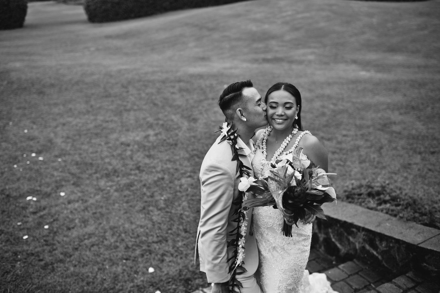 Groom kisses bride on cheek as she smiles holding bouquet, both wearing leis, black and white wedding photo.
