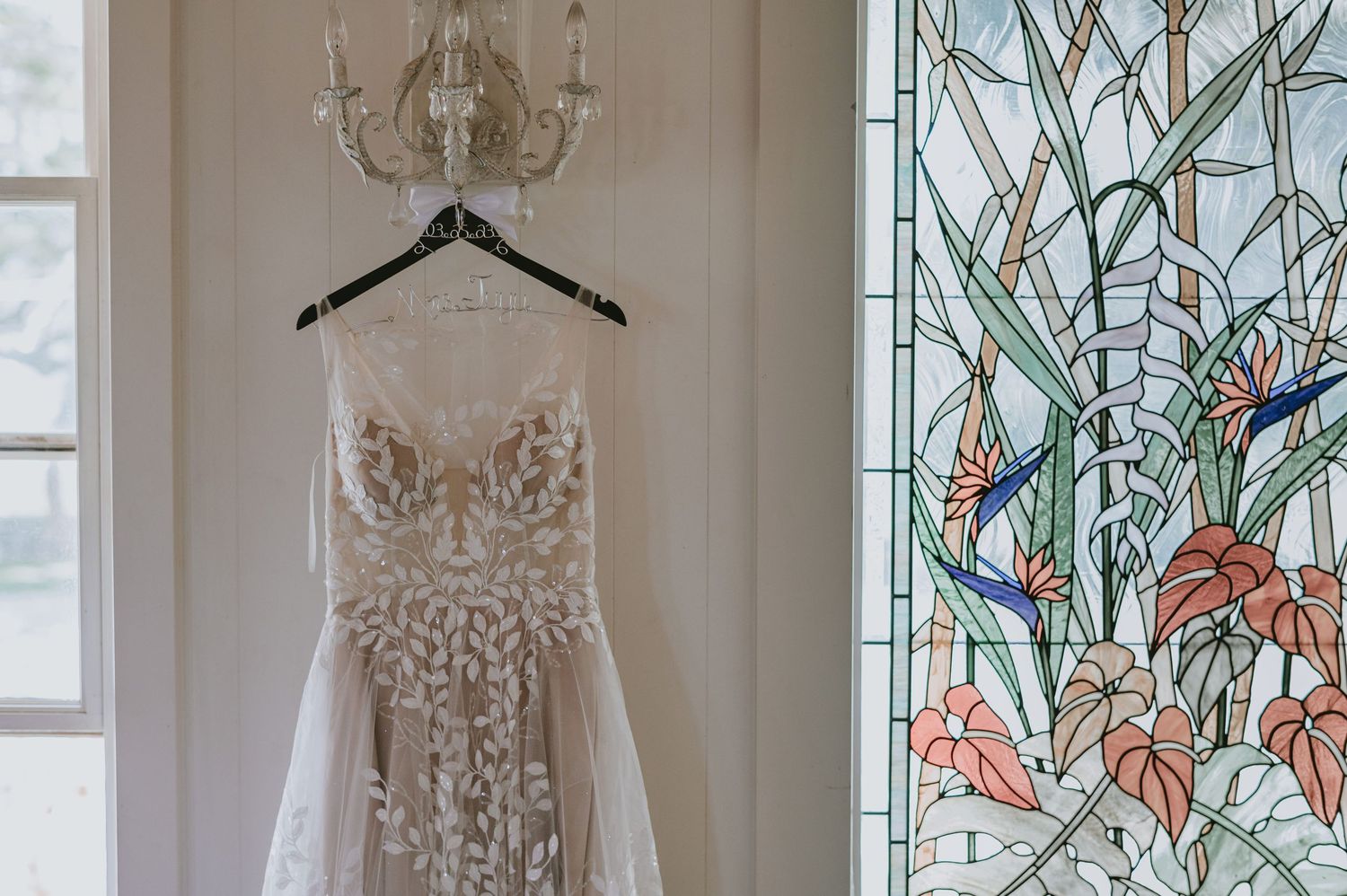 Intricate lace wedding dress hanging on chandelier beside colorful stained glass window indoors.
