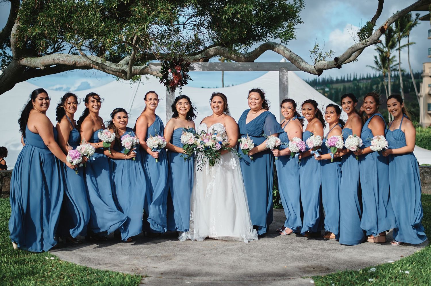 Large bridal party in blue dresses and formal wear poses for group photos on a sunny day.