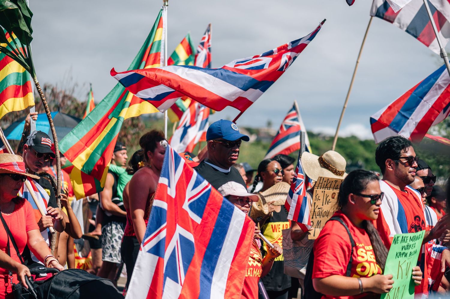 Crowd waves multiple Hawaiian state flags during daytime protest against development.