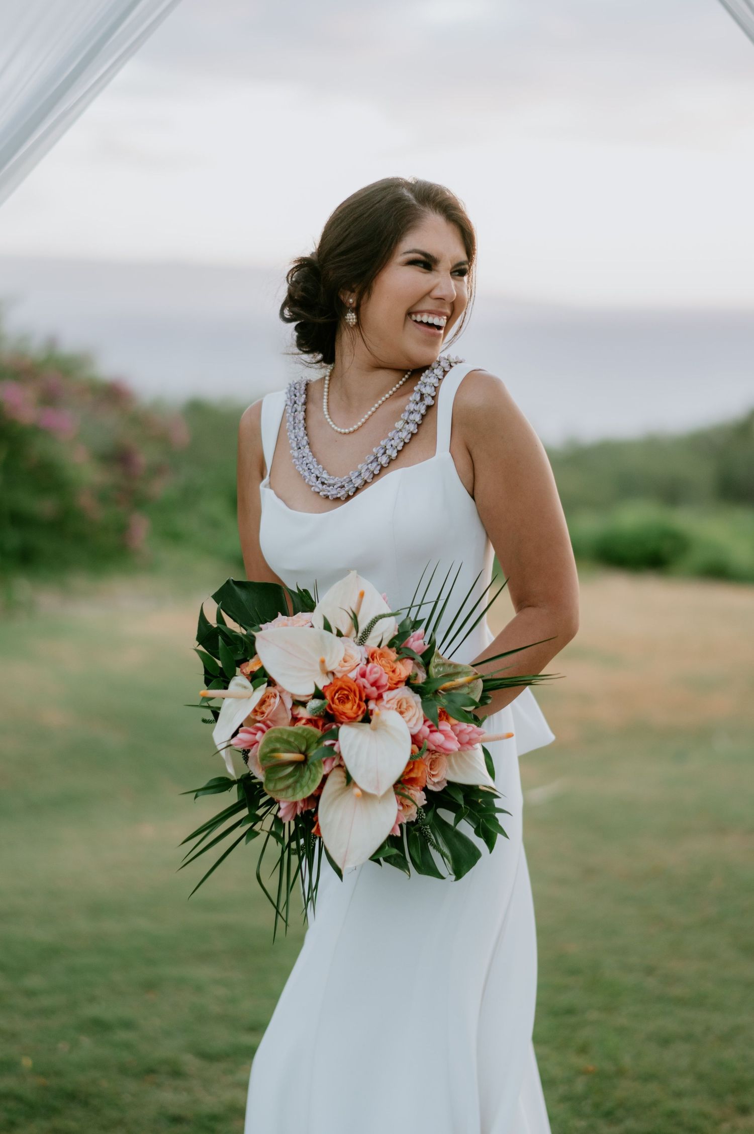 A beautiful bridal bouquet featuring coral and white flowers with tropical greenery against a grassy outdoor setting.