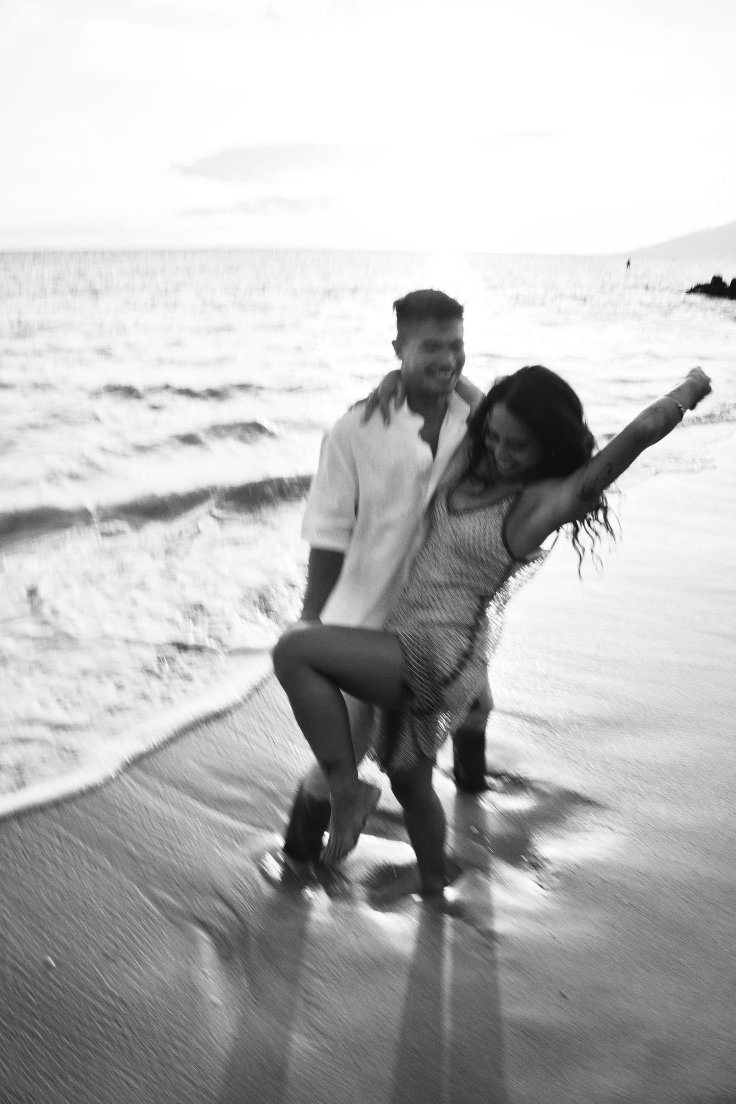 Joyful couple dancing in ocean waves on sandy beach at sunset, black and white.