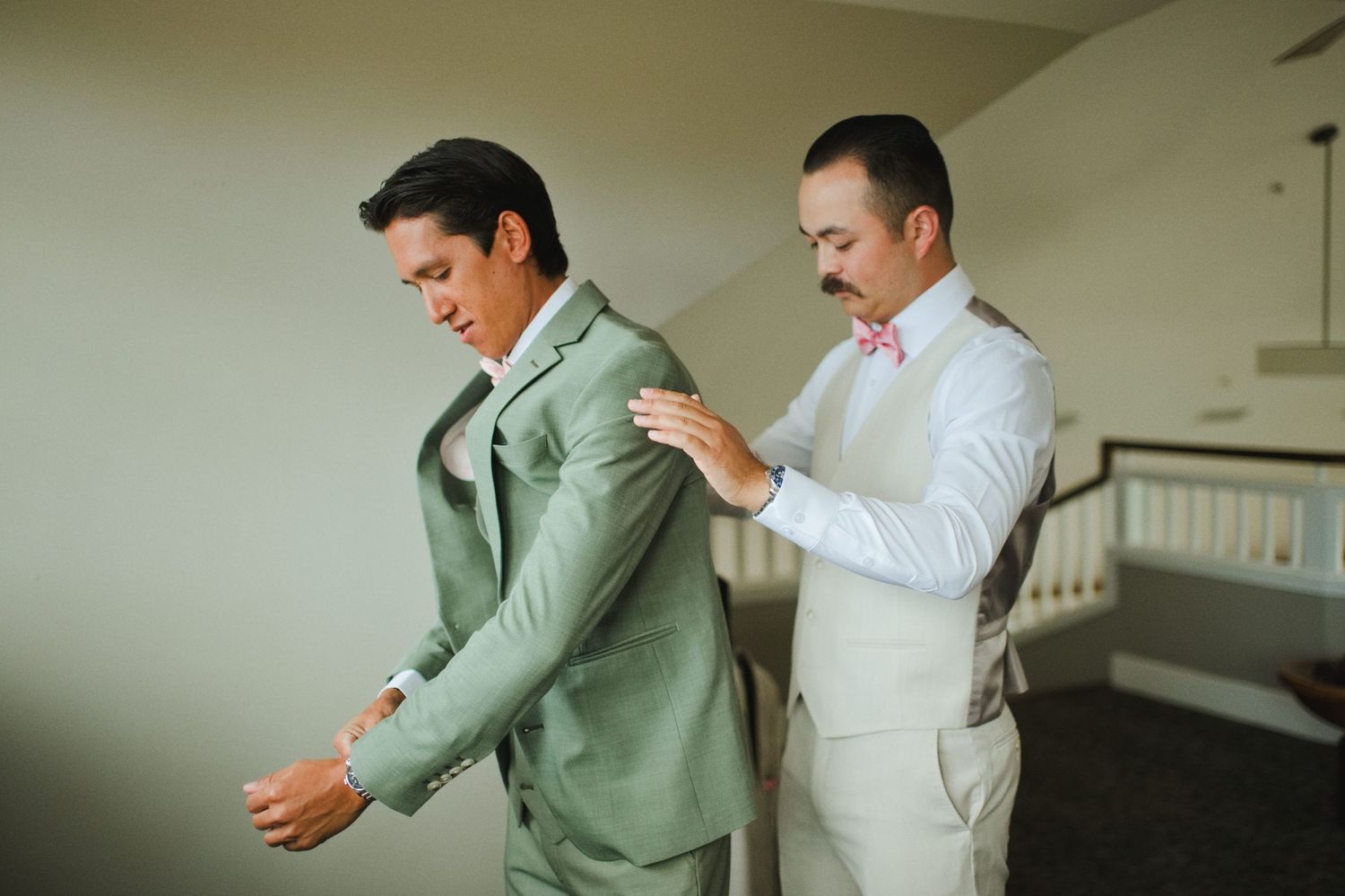 Groom in sage green suit gets jacket adjusted by groomsman in white shirt and pink bow tie before wedding ceremony.