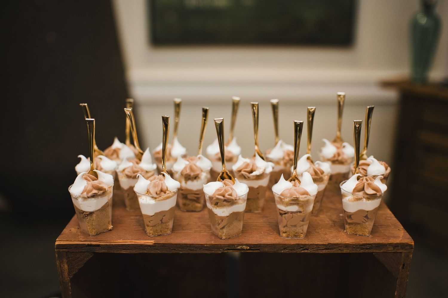 Elegant dessert cups with whipped cream and gold spoons arranged on a rustic wooden tray at a catered event.
