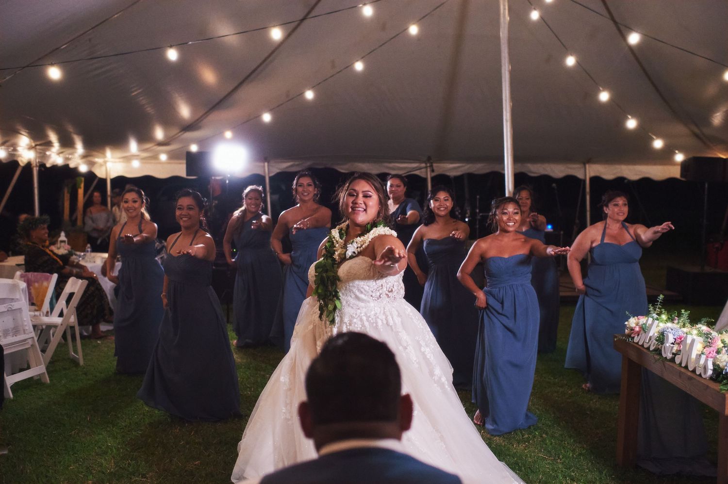Wedding party in blue dresses watches the newlyweds dance under a tent with string lights.