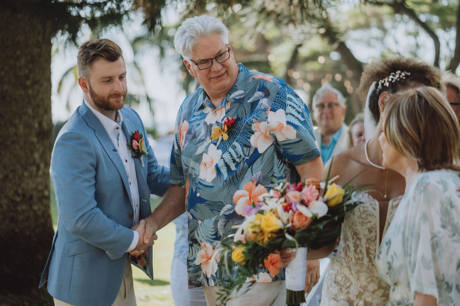 Wedding ceremony participants share emotional moments under tropical trees.