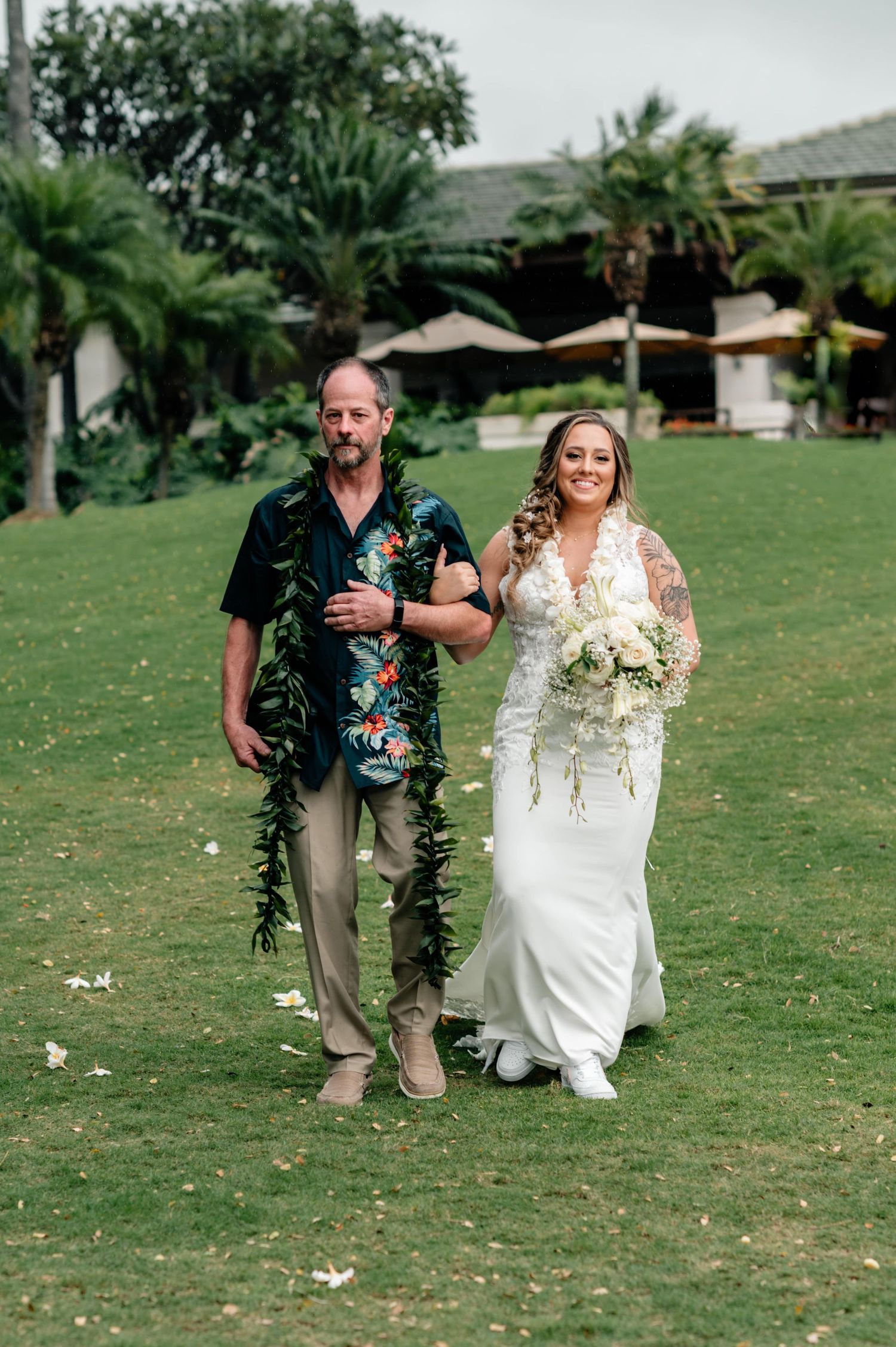 Wedding ceremony procession walking across green lawn with tropical foliage.