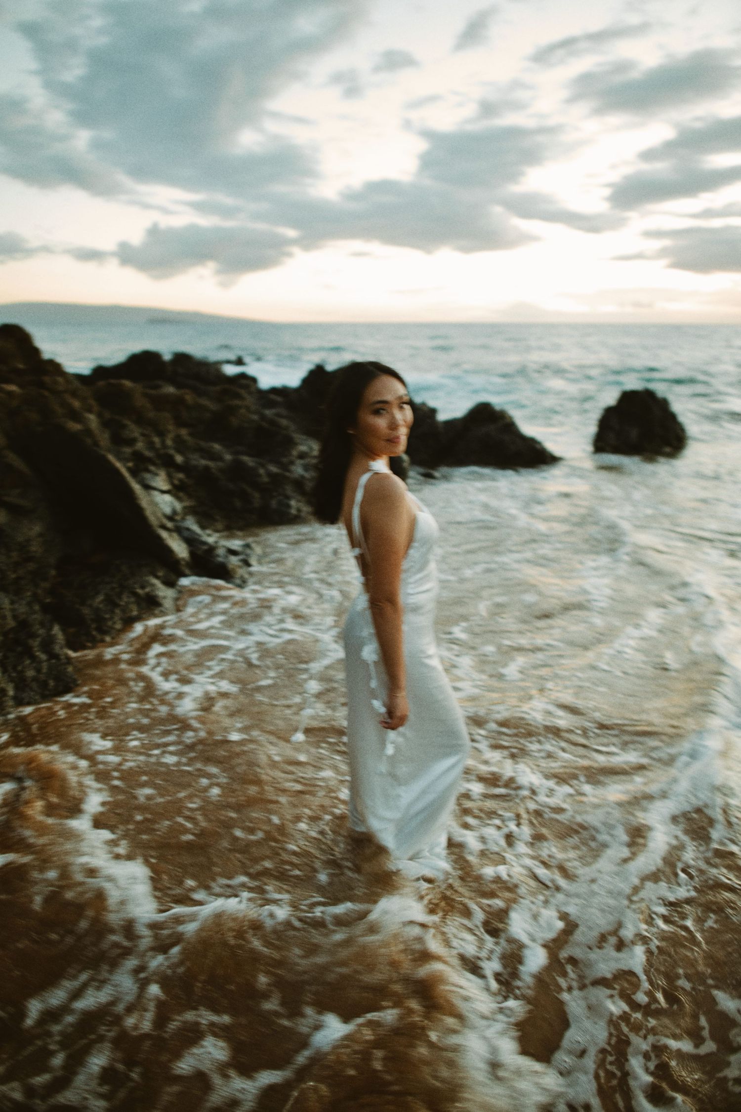 Woman in white dress standing in ocean waves on rocky beach at dramatic sunset.
