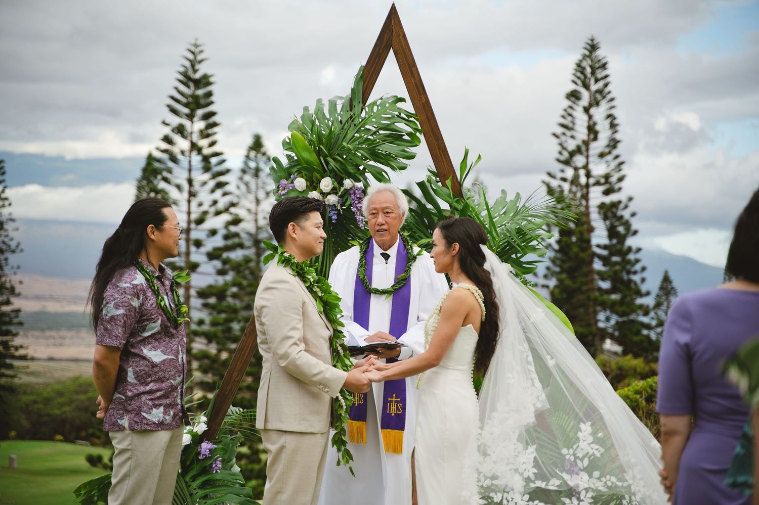Outdoor Hawaiian wedding ceremony with couple exchanging vows before officiant at wooden arch with tropical greenery.