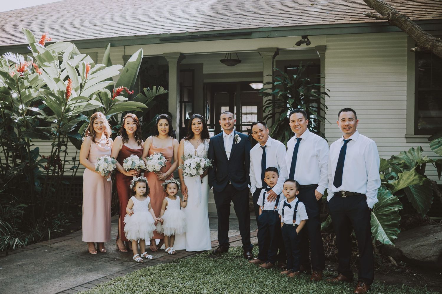 Wedding party posing outside a white house, bride and groom centered with bridesmaids, groomsmen, and children in formal attire.