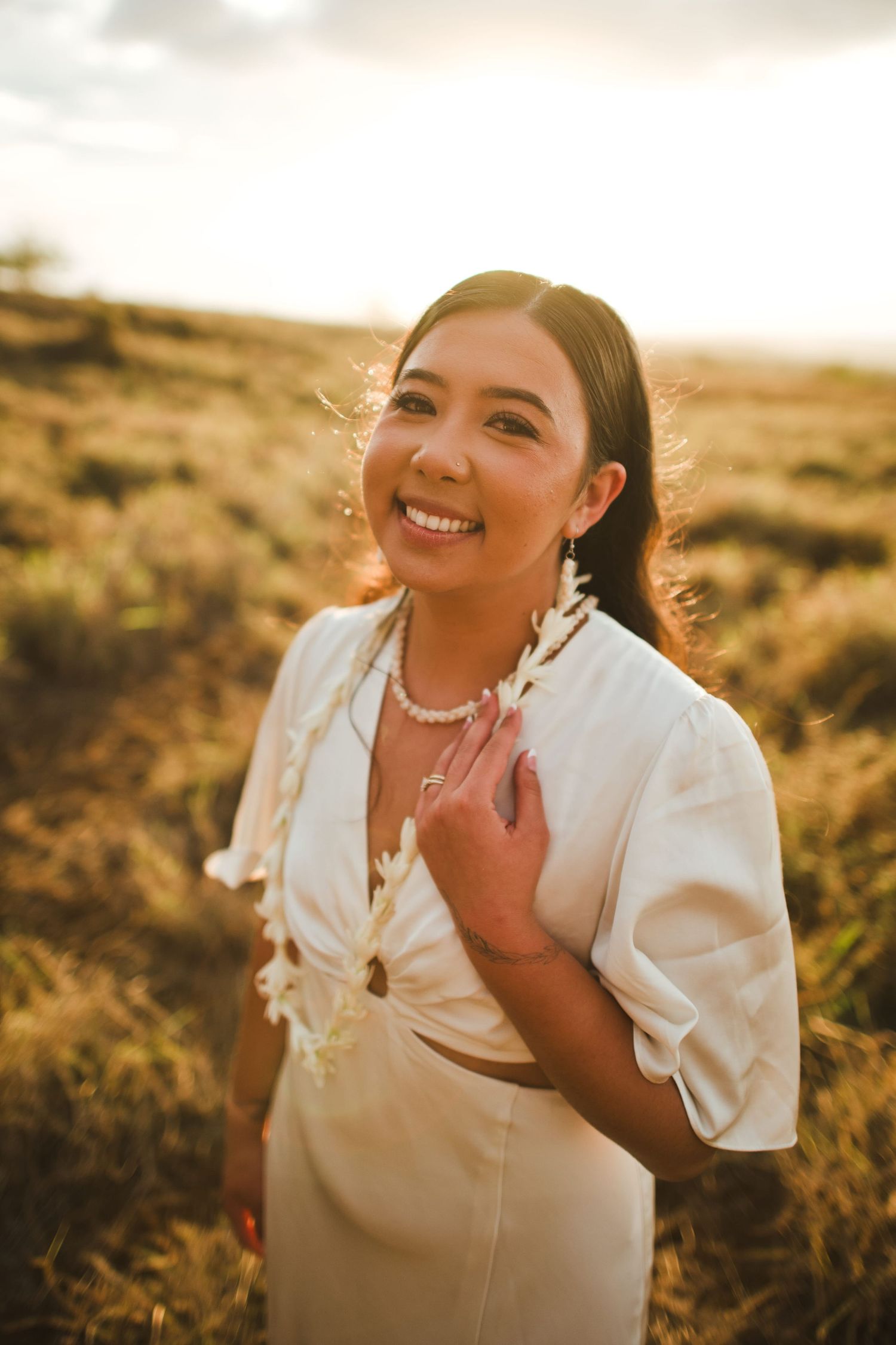 Smiling woman in white outfit wearing a floral lei, posing in a golden field during sunset.