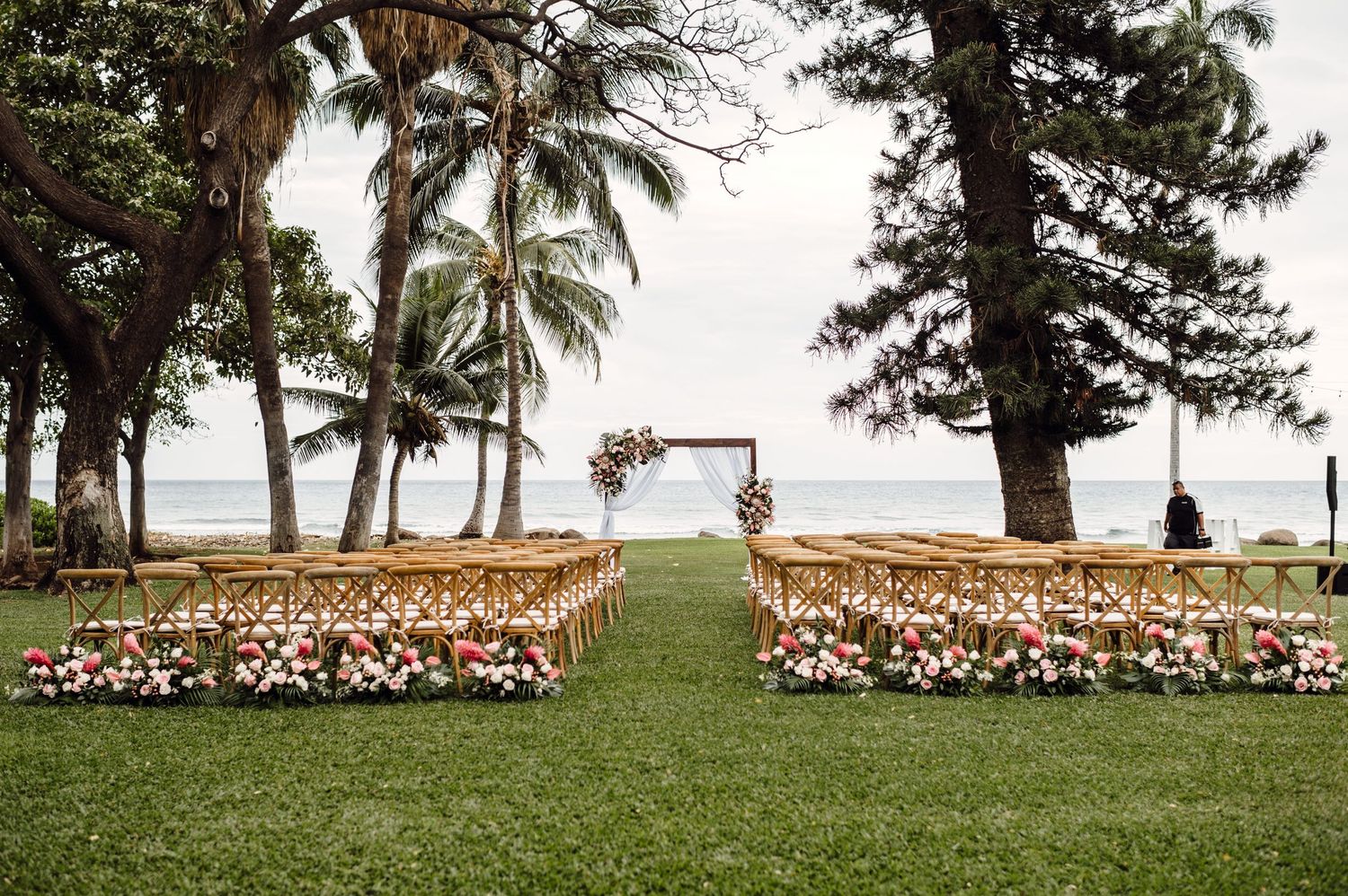 Outdoor wedding ceremony setup on green lawn with floral-lined aisle, wooden chairs, arch, and ocean view framed by palm trees.