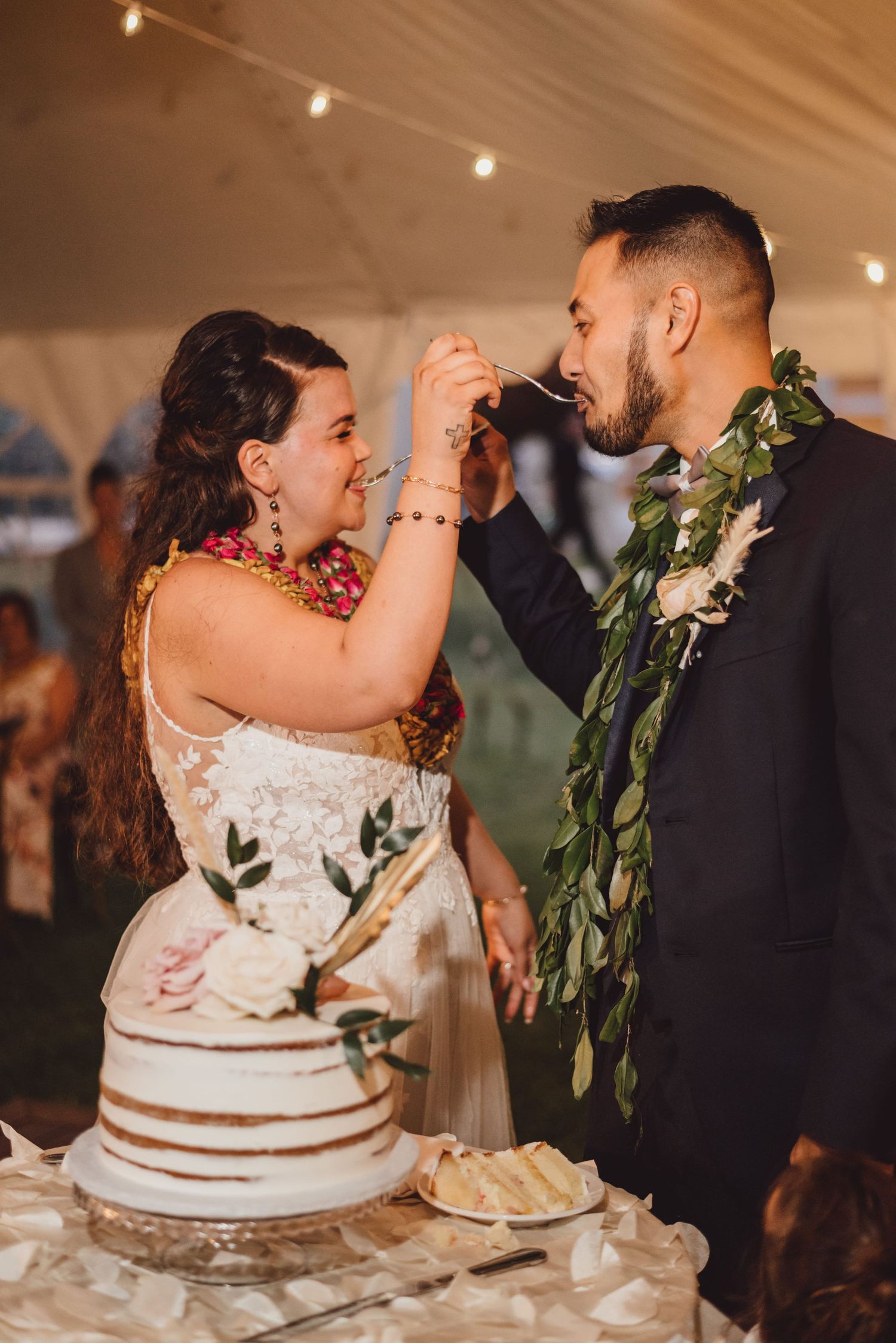 Bride feeds groom wedding cake under string lights at Hawaiian-themed reception, both wearing leis.