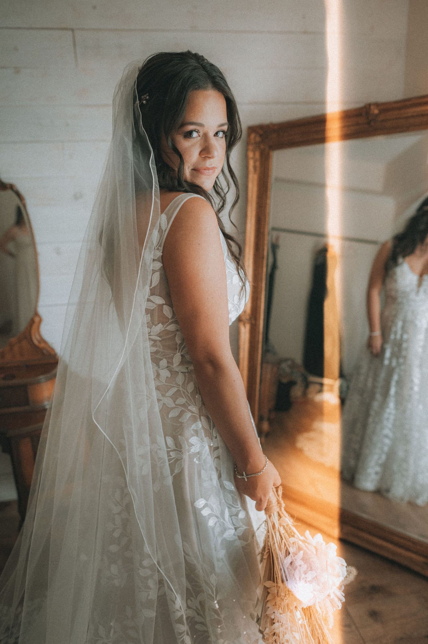 Bride in lace wedding gown and veil holds bouquet, glancing over shoulder near gold mirror in warm sunlit room.