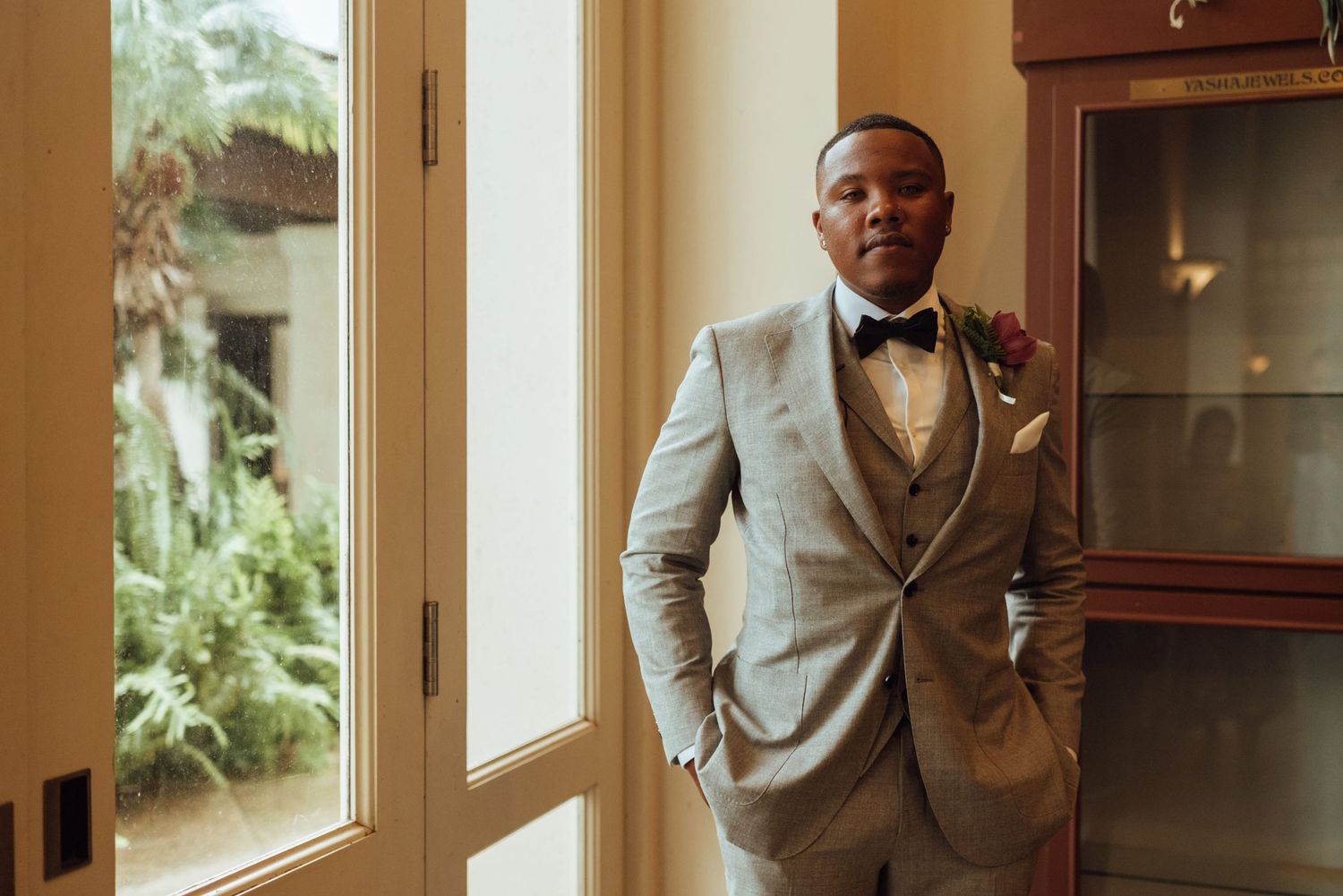 A well-dressed groom in a light gray suit and bow tie poses confidently near a window with tropical greenery outside.