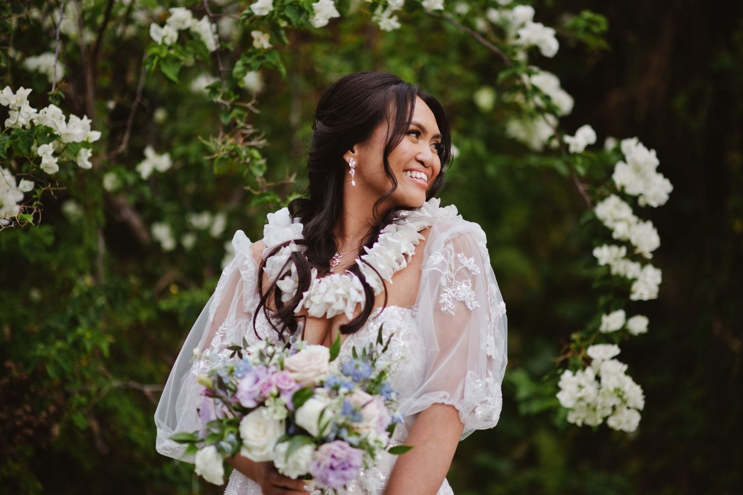 Smiling bride in lace gown holds floral bouquet surrounded by white blooming flowers outdoors.