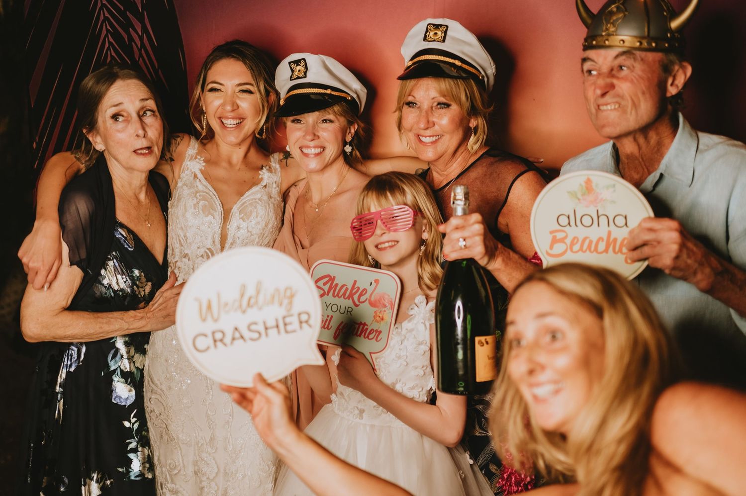 Group of wedding guests posing in photo booth with fun props including Wedding Crasher and Aloha Beaches signs.
