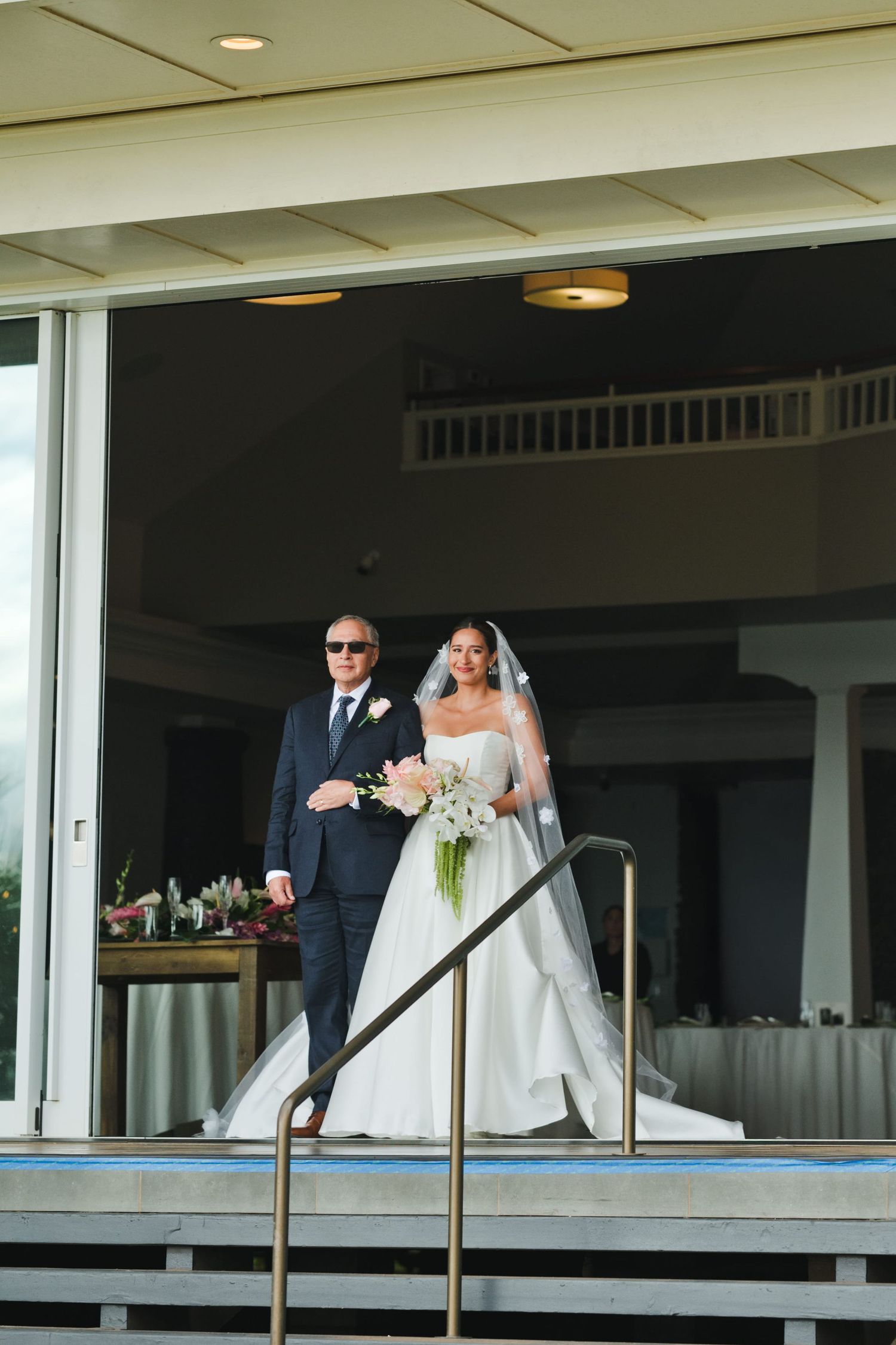 Bride in white gown and veil walks down steps with escort in dark suit at elegant wedding venue.