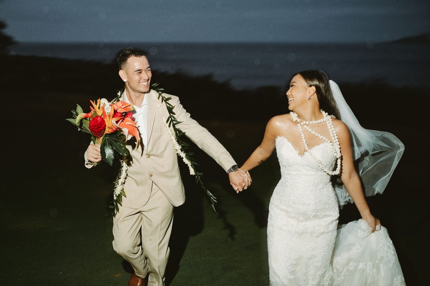 Smiling newlyweds walking hand in hand at dusk with a tropical floral bouquet in Hawaii.