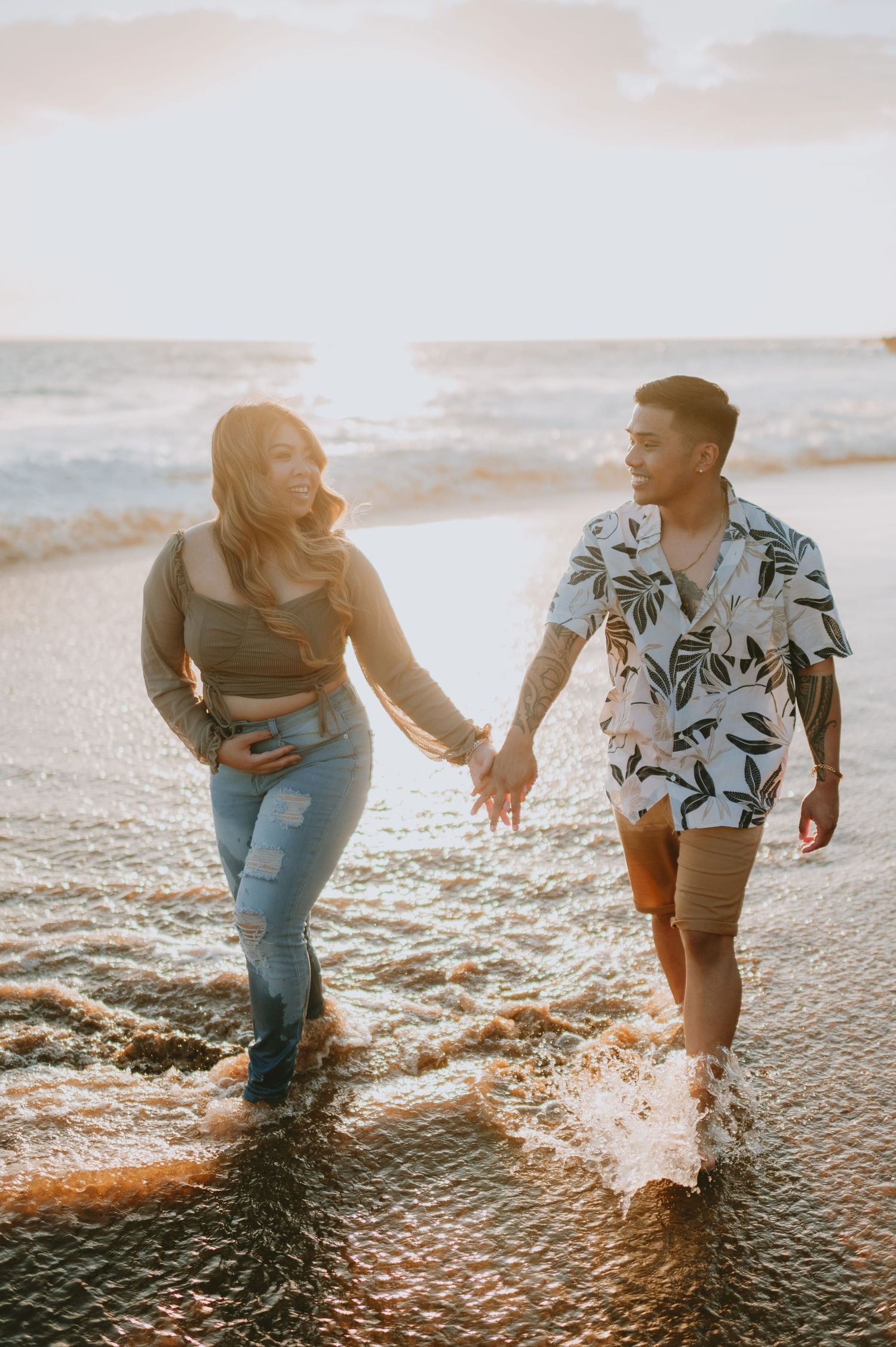 Couple laughing and holding hands while walking through ocean waves at sunset.