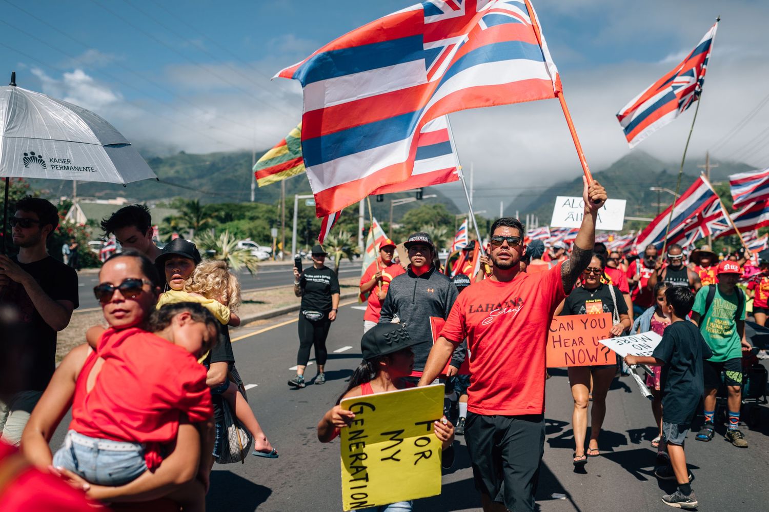 Crowd of protesters in red shirts wave flags and hold signs during a Hawaiian demonstration.