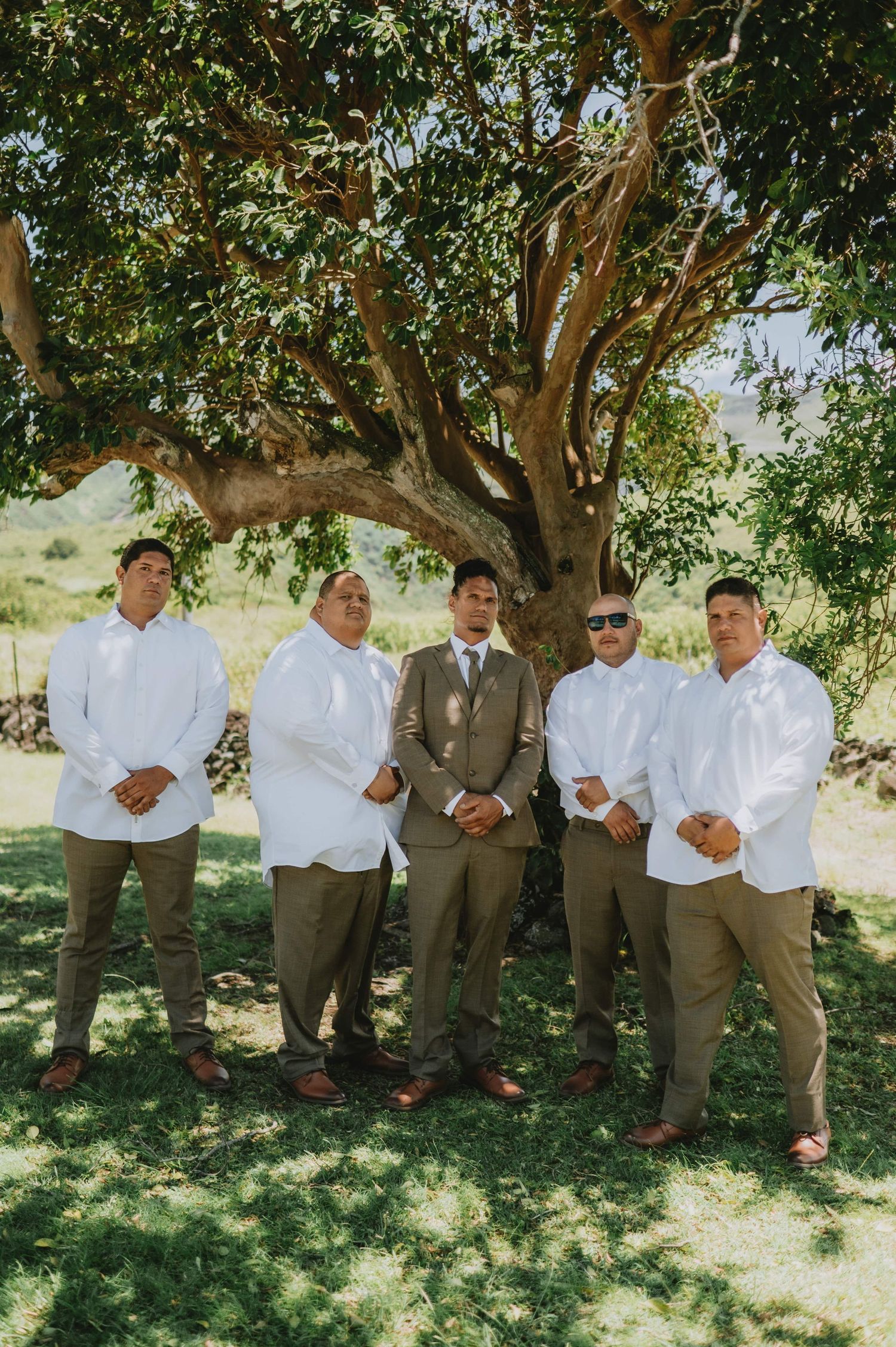 Wedding party members in white shirts and khaki pants pose casually under a shady tree.