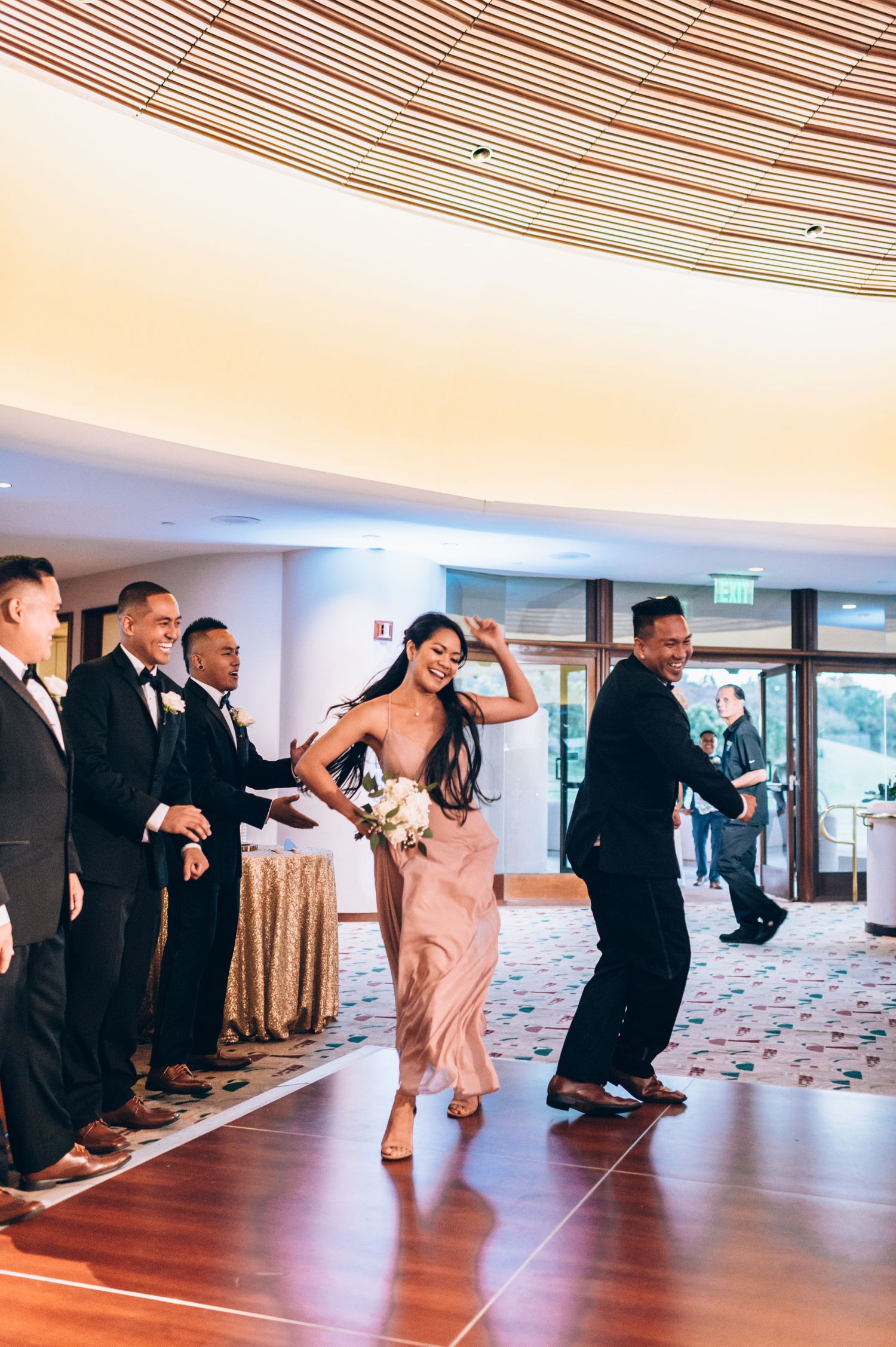 Wedding party performs energetic dance routine in formal attire against curved ceiling backdrop.