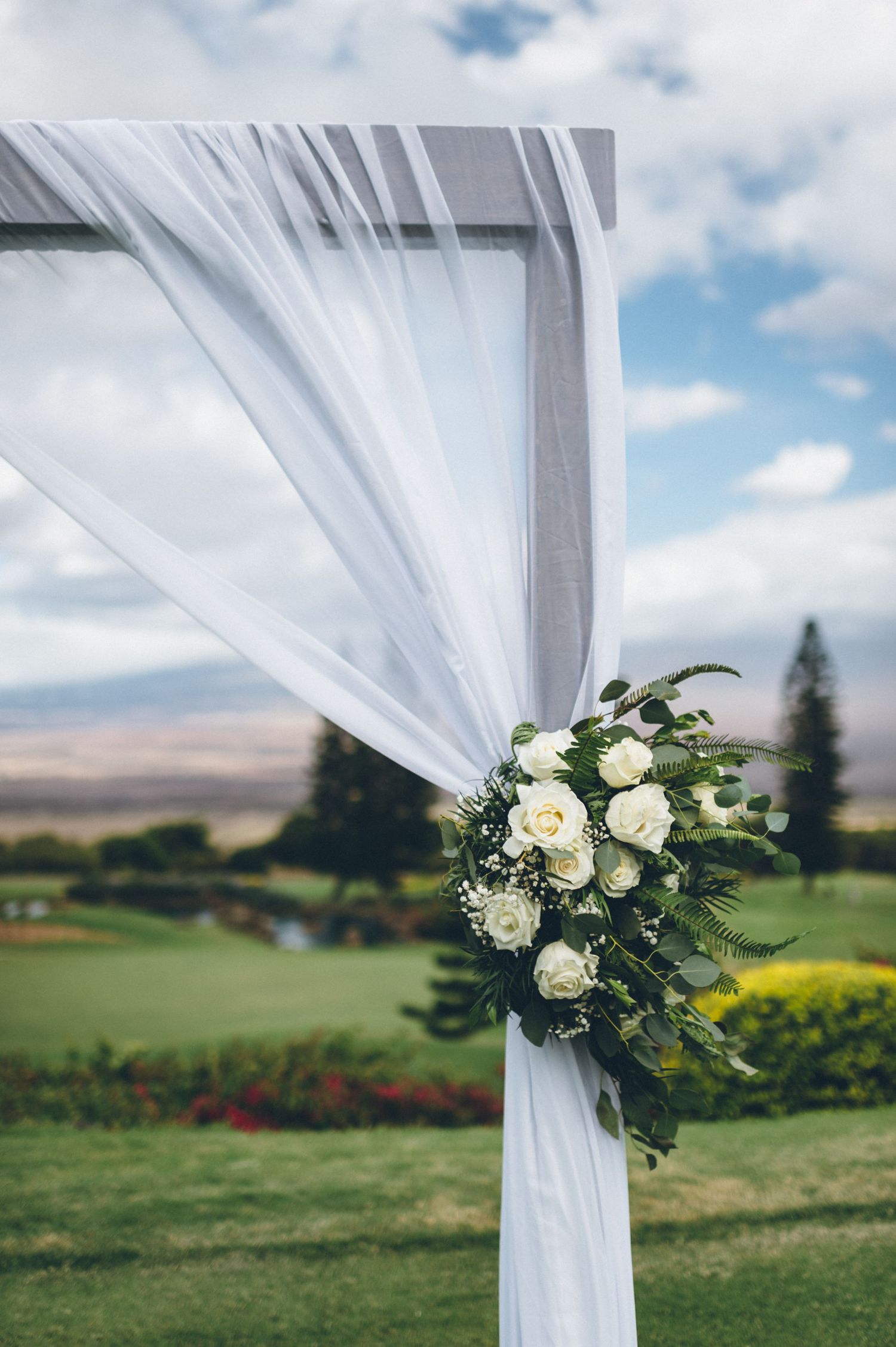 White wedding arch decorated with elegant white roses and greenery against a scenic mountain backdrop.