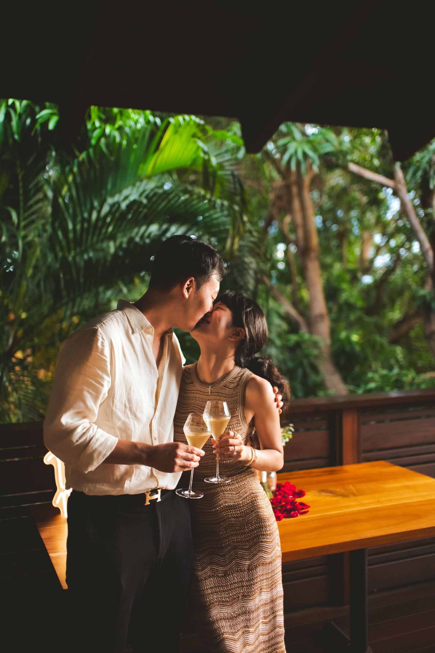 Couple sharing a romantic kiss while holding champagne glasses on a tropical outdoor terrace surrounded by lush greenery.
