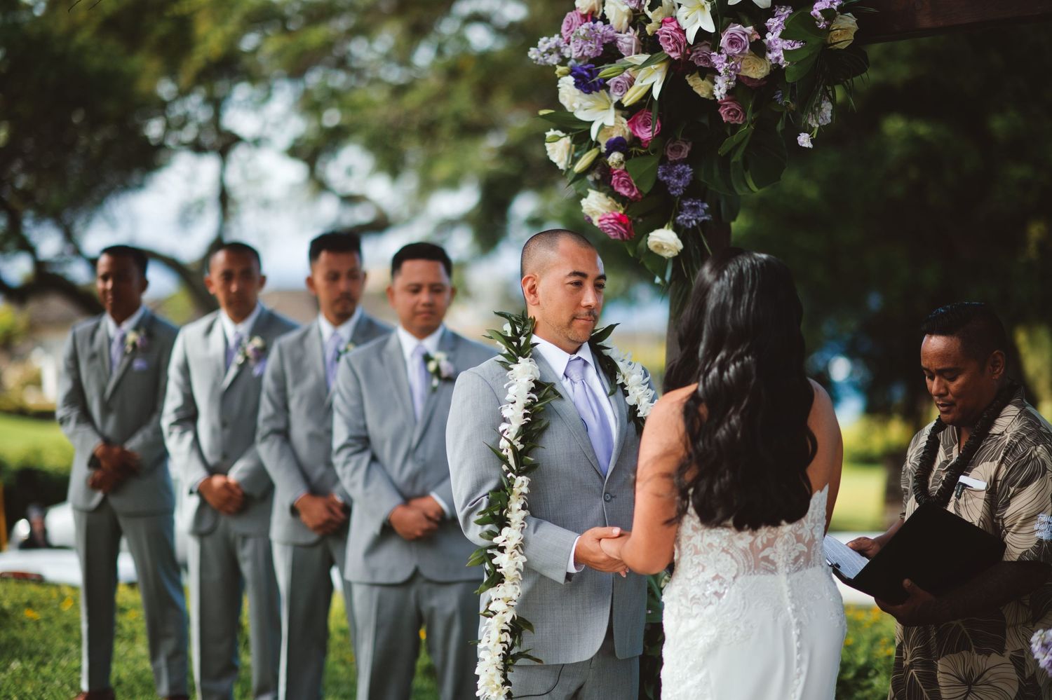 Outdoor wedding ceremony with groom in gray suit holding leis, bride in white dress, groomsmen, and floral arch.